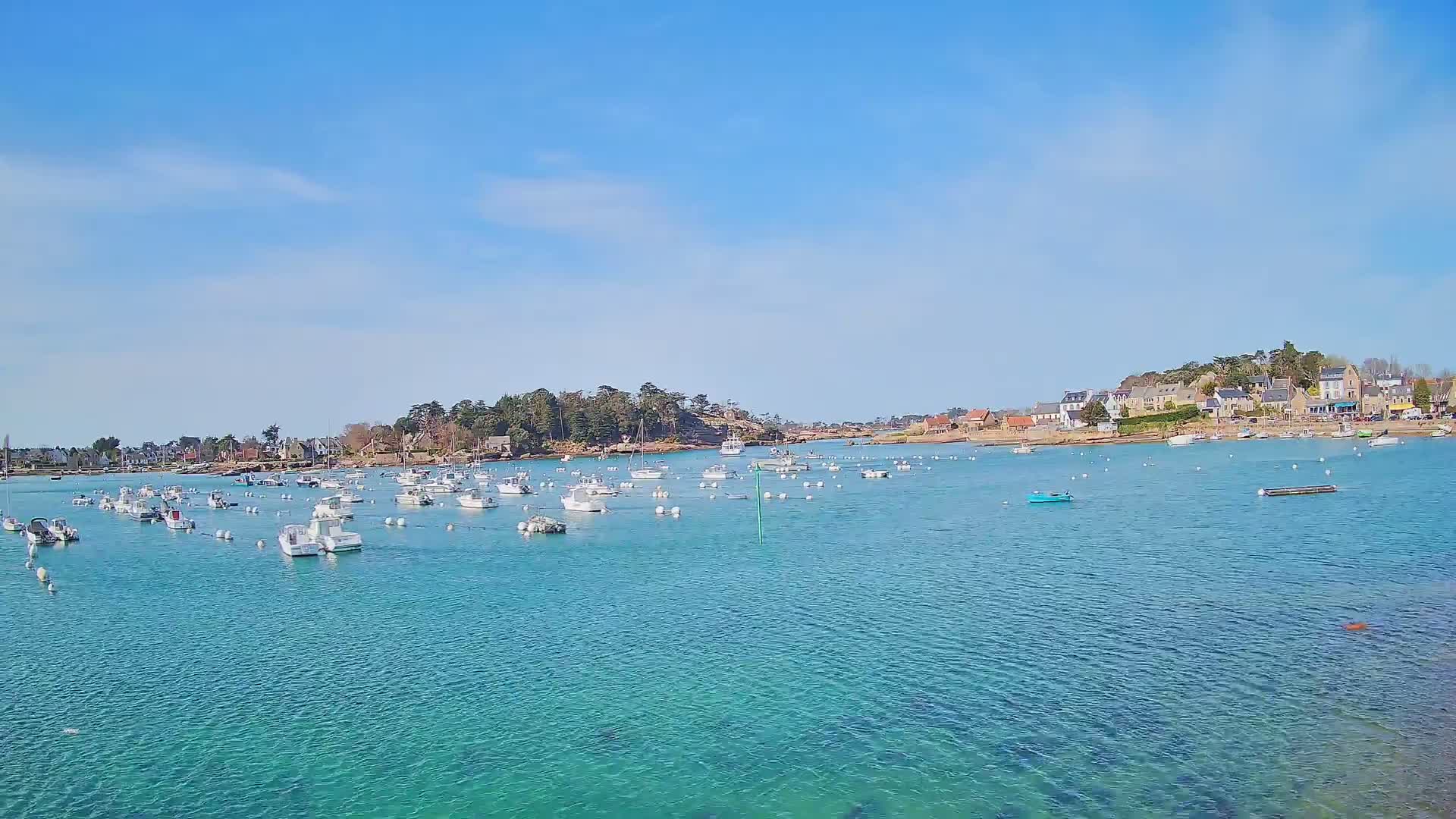 Numerous small white boats are moored in a calm, clear turquoise body of water, with a small village situated on a shoreline in the background under a bright, sunny sky with a few scattered clouds.