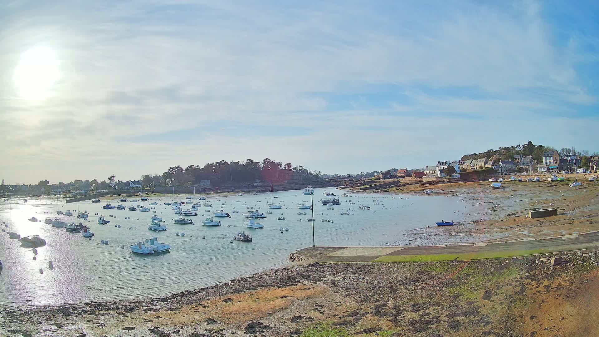 A calm harbor filled with numerous small boats sits partly exposed at low tide under a mostly sunny sky.