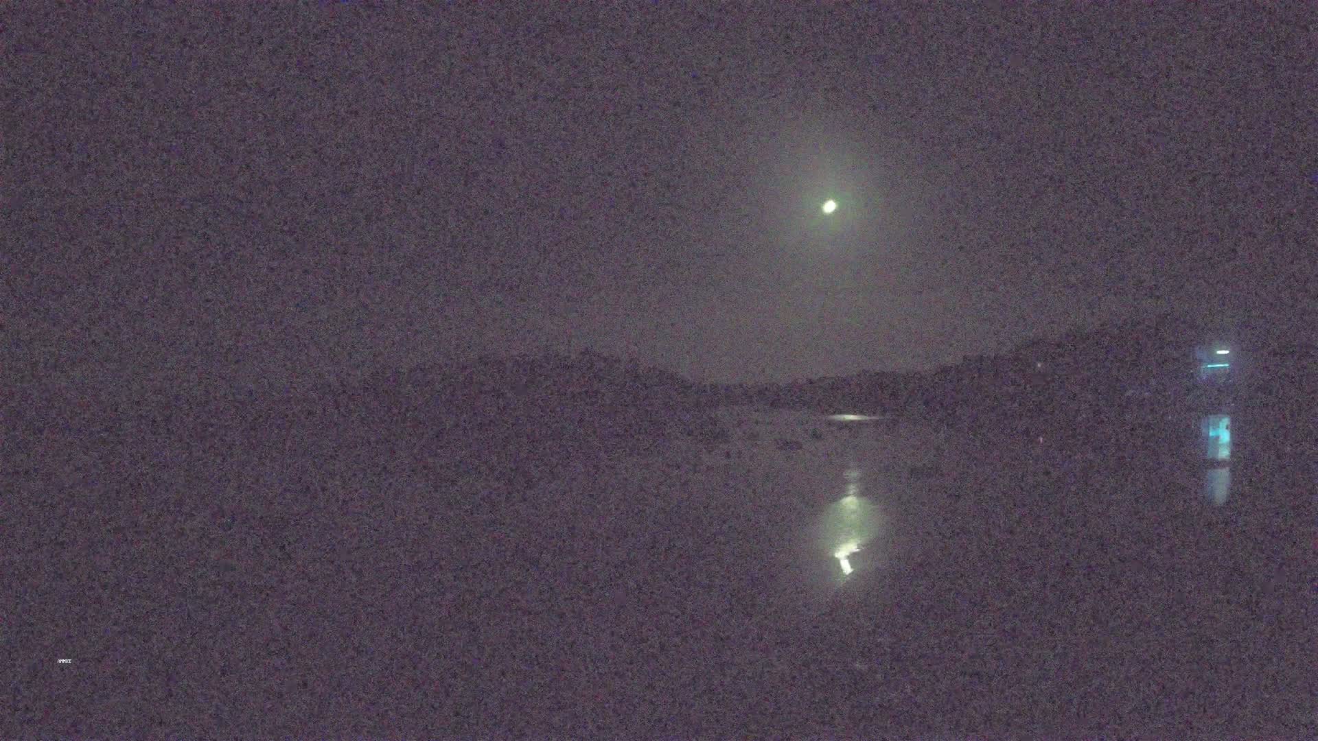 A bright moon illuminates a dark, still body of water reflecting lights from the opposite shore under a night sky.