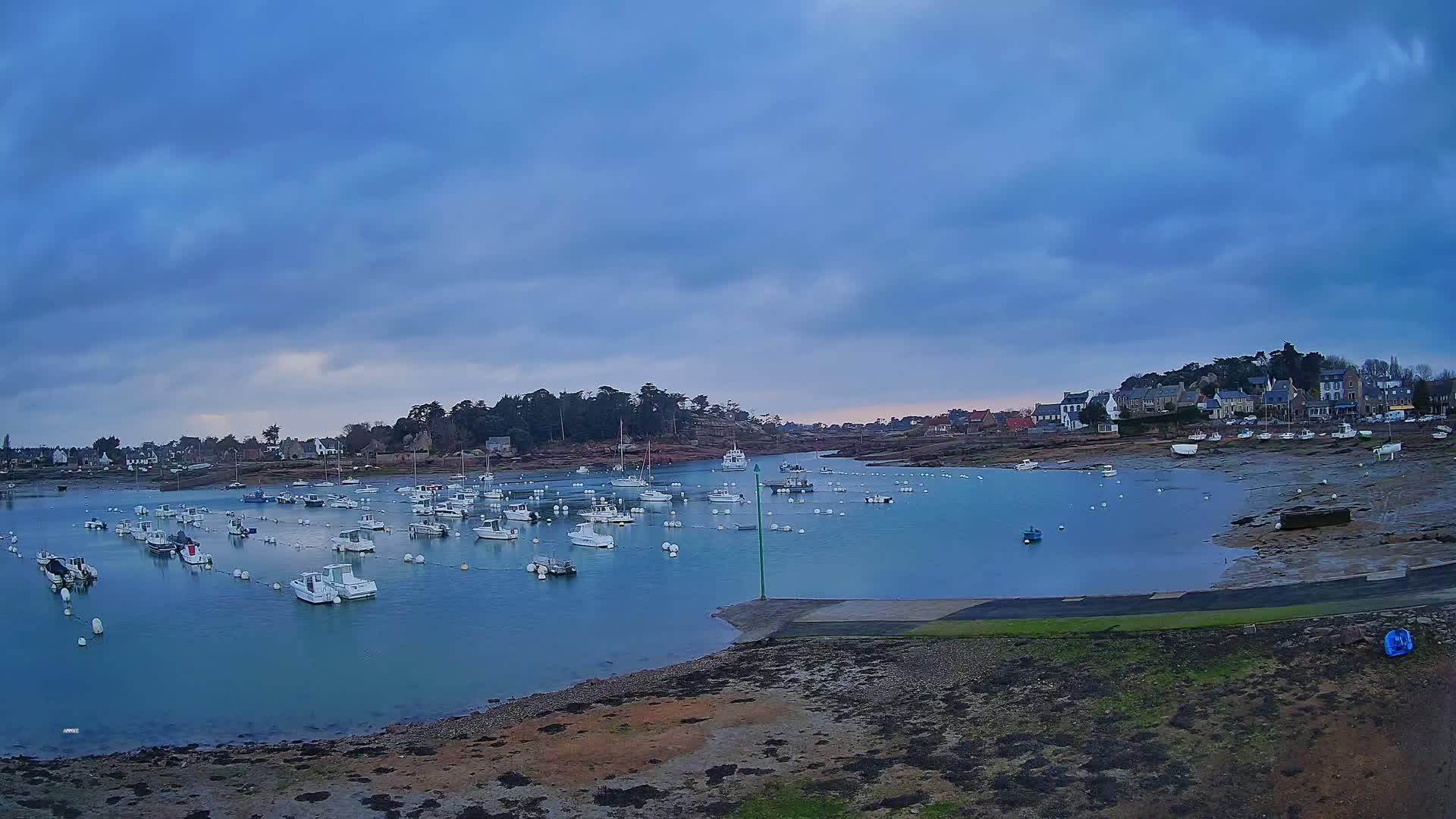 Many small white boats are moored in a calm, blue harbor under a cloudy, twilight sky, with a small village visible in the background.