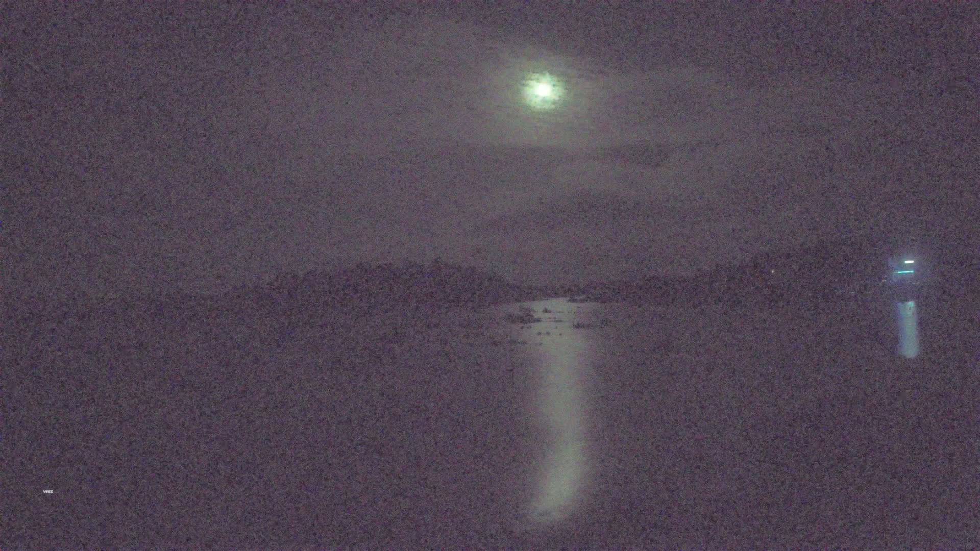 A moonlit night scene shows a body of water reflecting light, with dark land visible in the distance under a mostly cloudy sky.