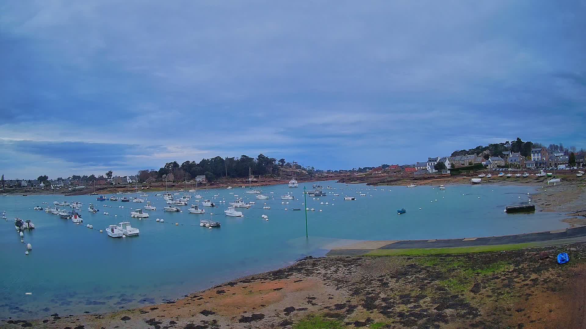 Numerous small boats are moored in a calm, teal-colored bay under a cloudy sky, with a small village visible on the opposite shore.