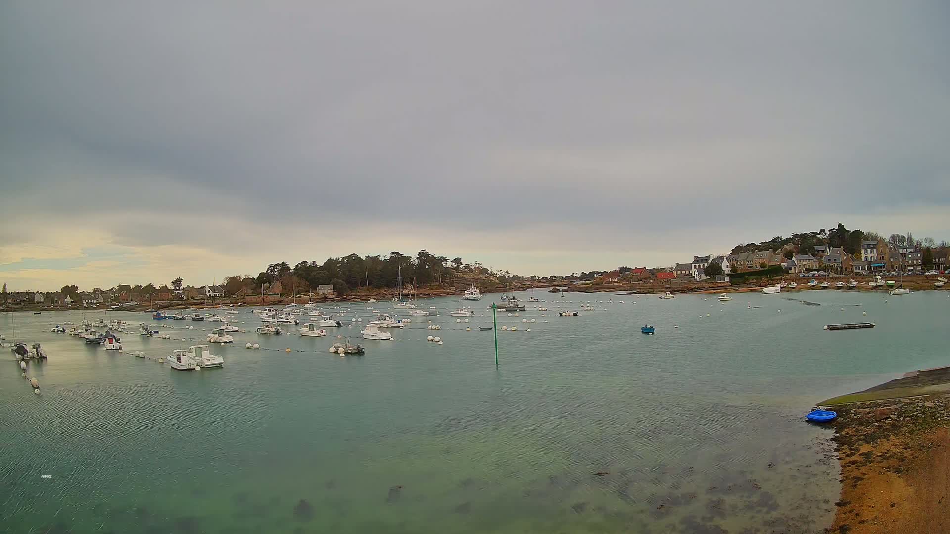 Many small boats are moored in a calm, teal-colored harbor under an overcast sky, near a small town nestled on a rocky shoreline.
