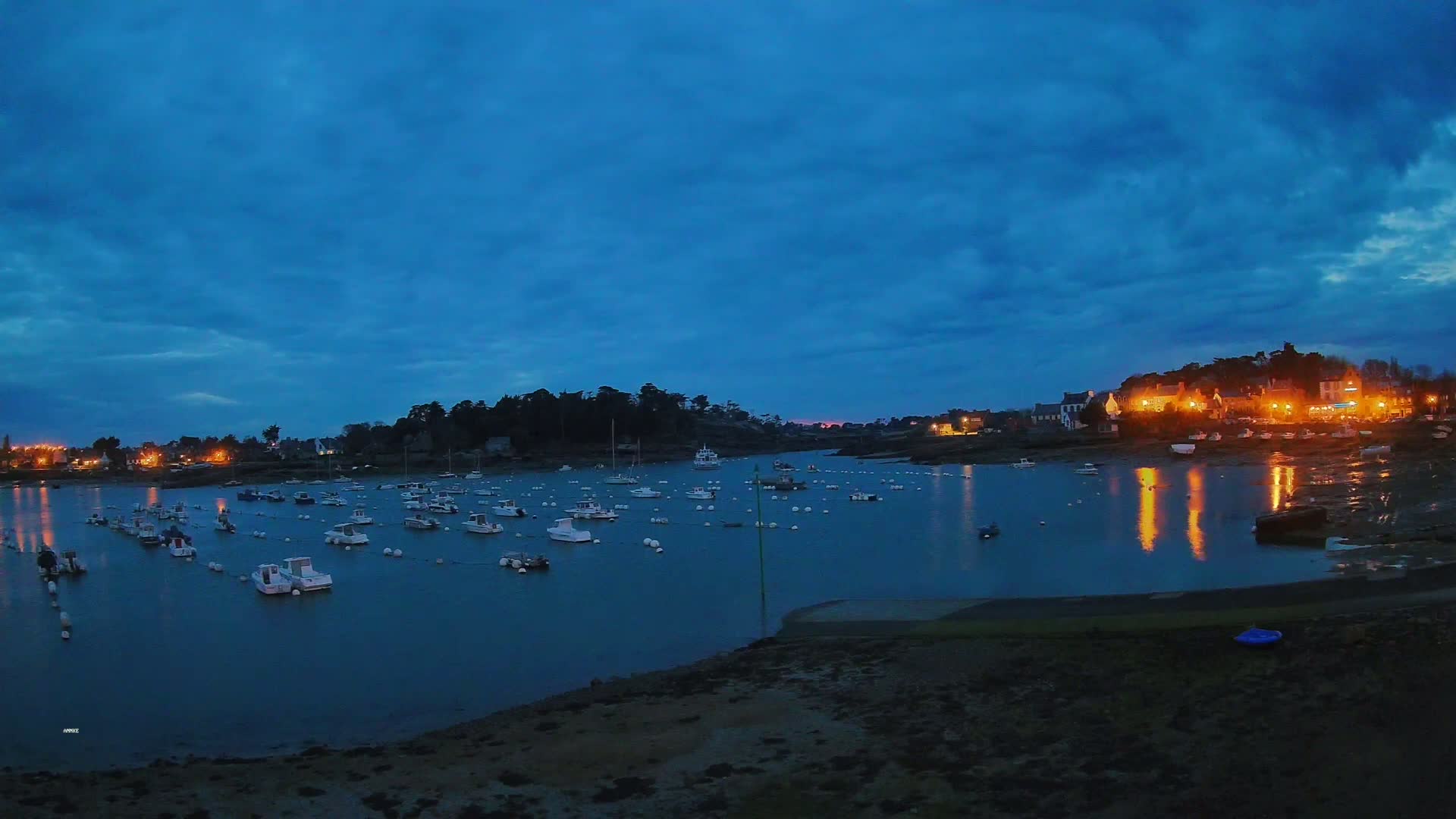 Many small boats are moored in a calm harbor at twilight under a mostly cloudy, dark blue sky, with a village's lights reflecting on the water in the background.