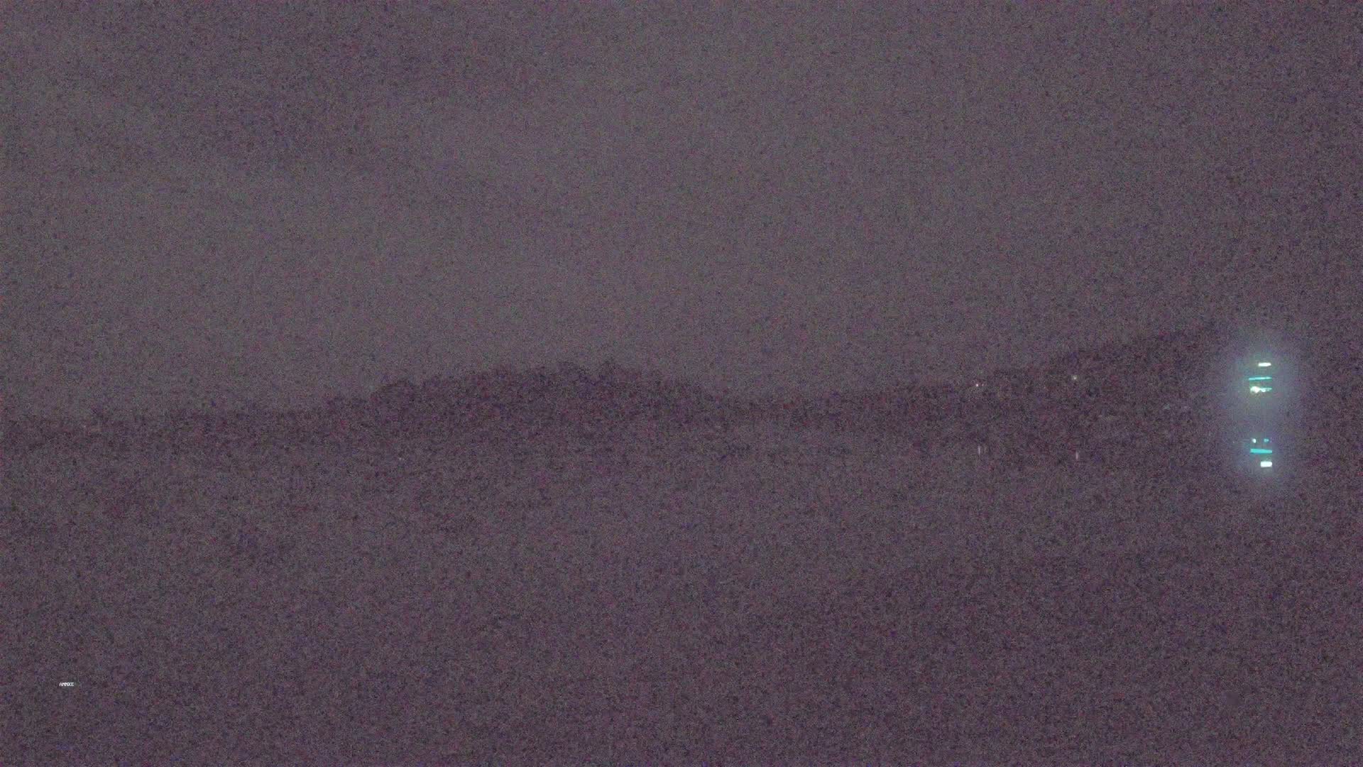 A dimly lit, dark landscape is visible under a gray, overcast sky, with a tall structure faintly illuminated on the right.