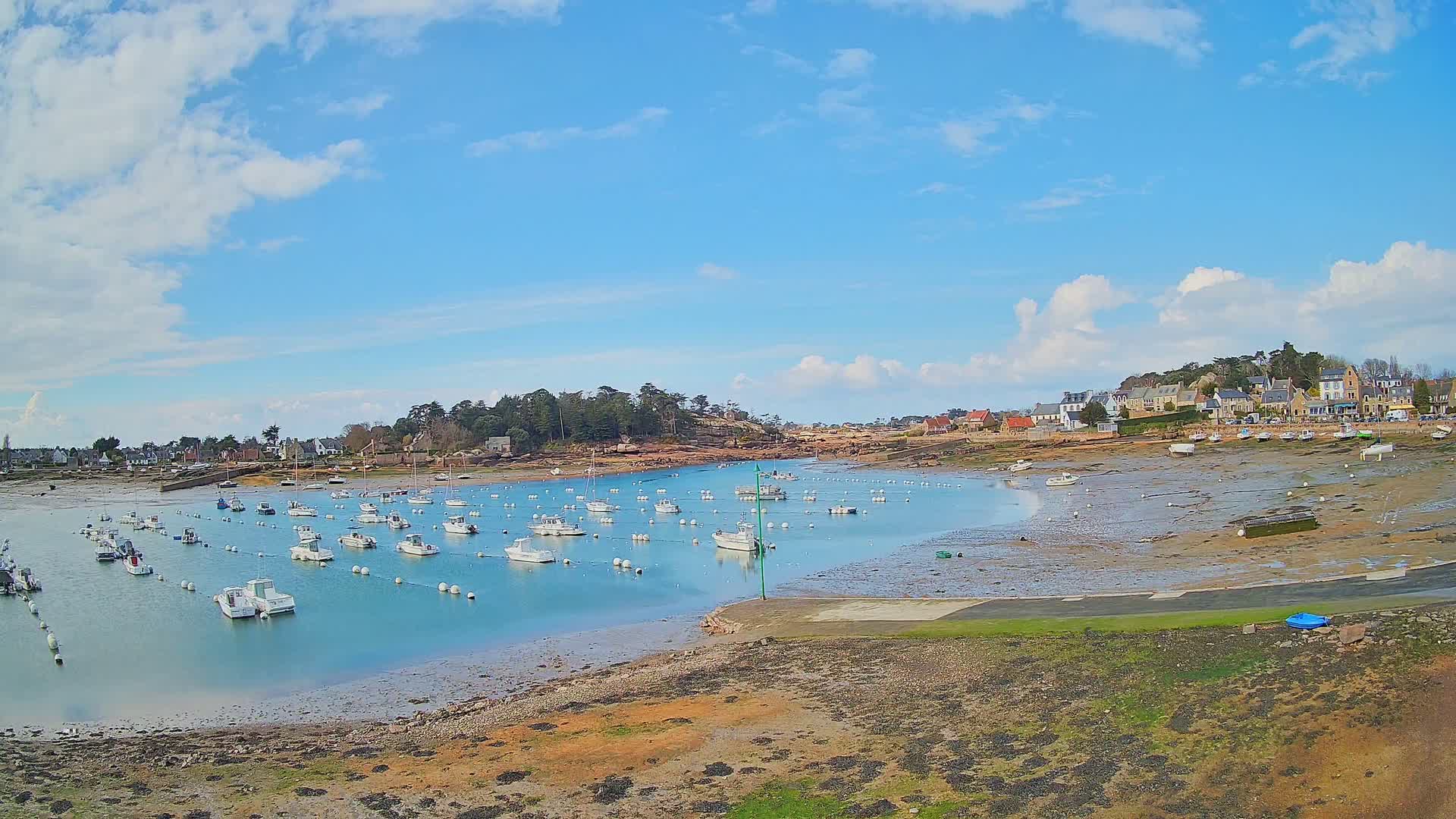 Numerous small white boats are moored in a calm bay under a partly cloudy blue sky, with a small village visible on the shore.