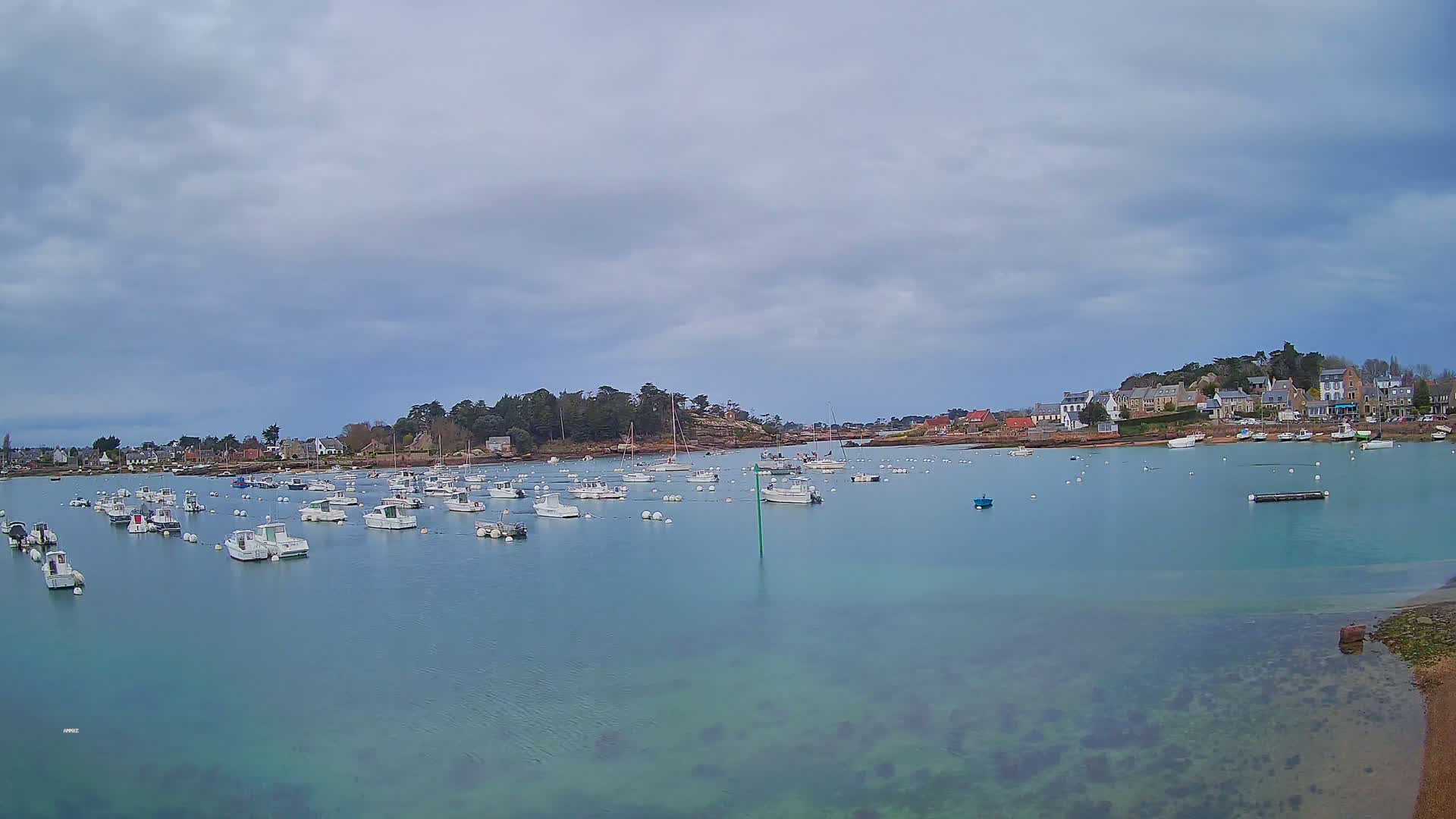 Many small white boats are moored in a calm, light teal bay under a cloudy sky, with a small village visible on the far shore.