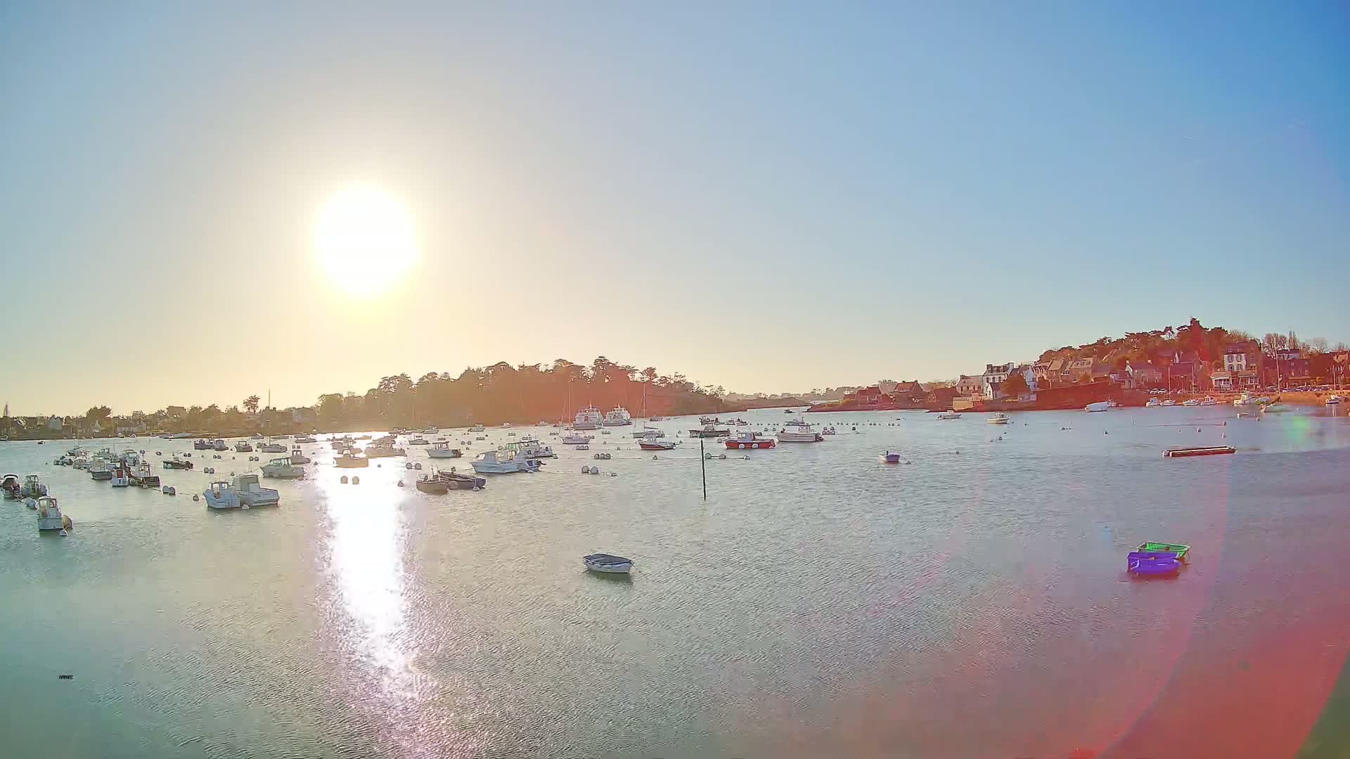 Many small boats are moored in a calm harbor under a bright, sunny sky, with a village visible in the background.