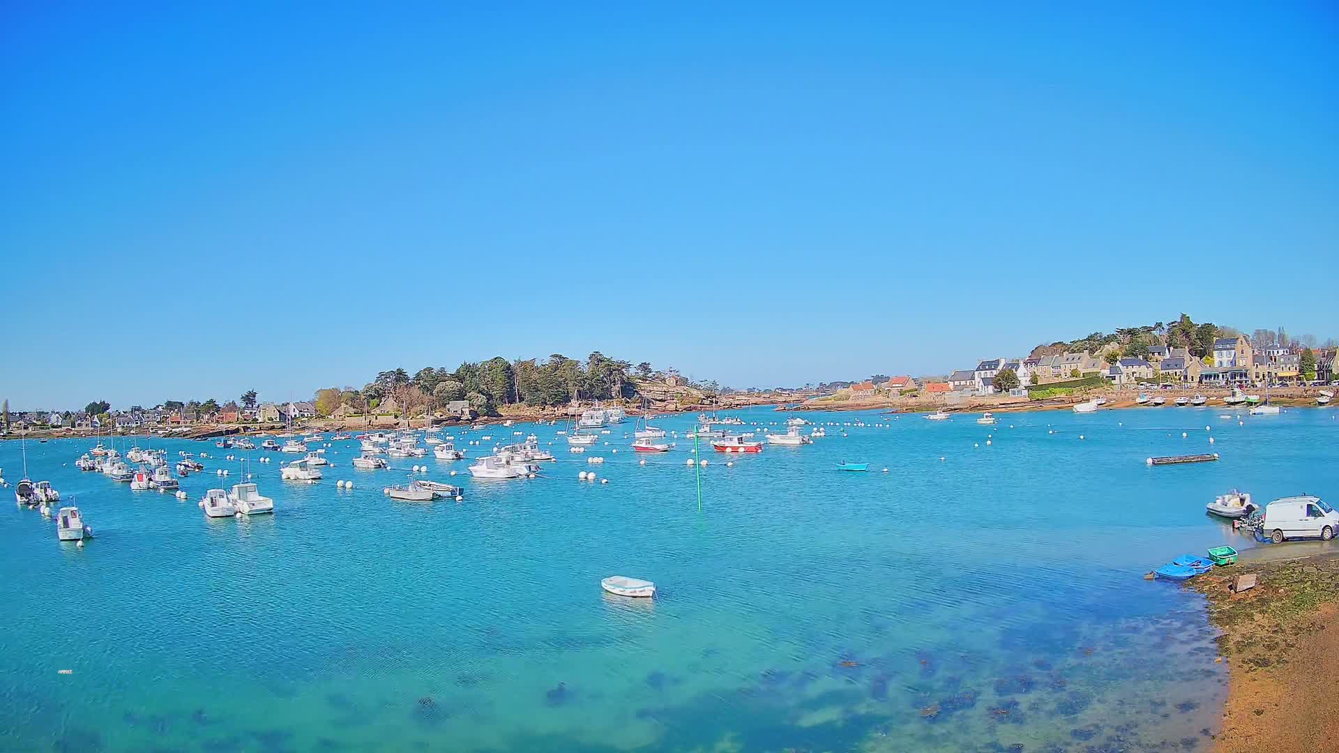 A harbor filled with numerous small boats under a clear blue sky, surrounded by a calm, turquoise sea and a shoreline with buildings and trees.