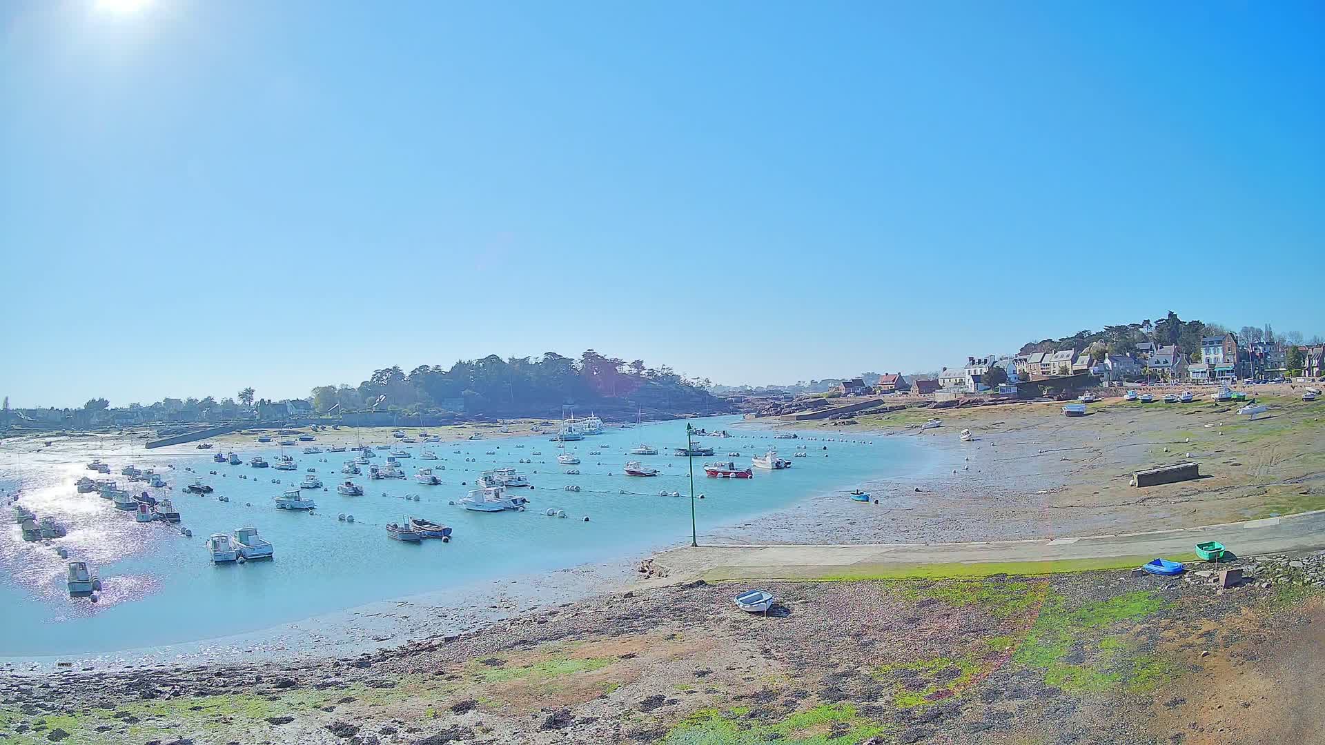 Under a bright, sunny sky, numerous small boats are moored in a calm harbor near a small village, with exposed mudflats visible at low tide.