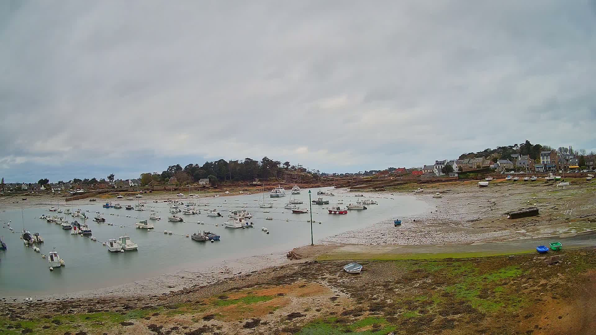 Many small boats are moored in a calm harbor at low tide under a cloudy sky, near a small village nestled beside a rocky shoreline.