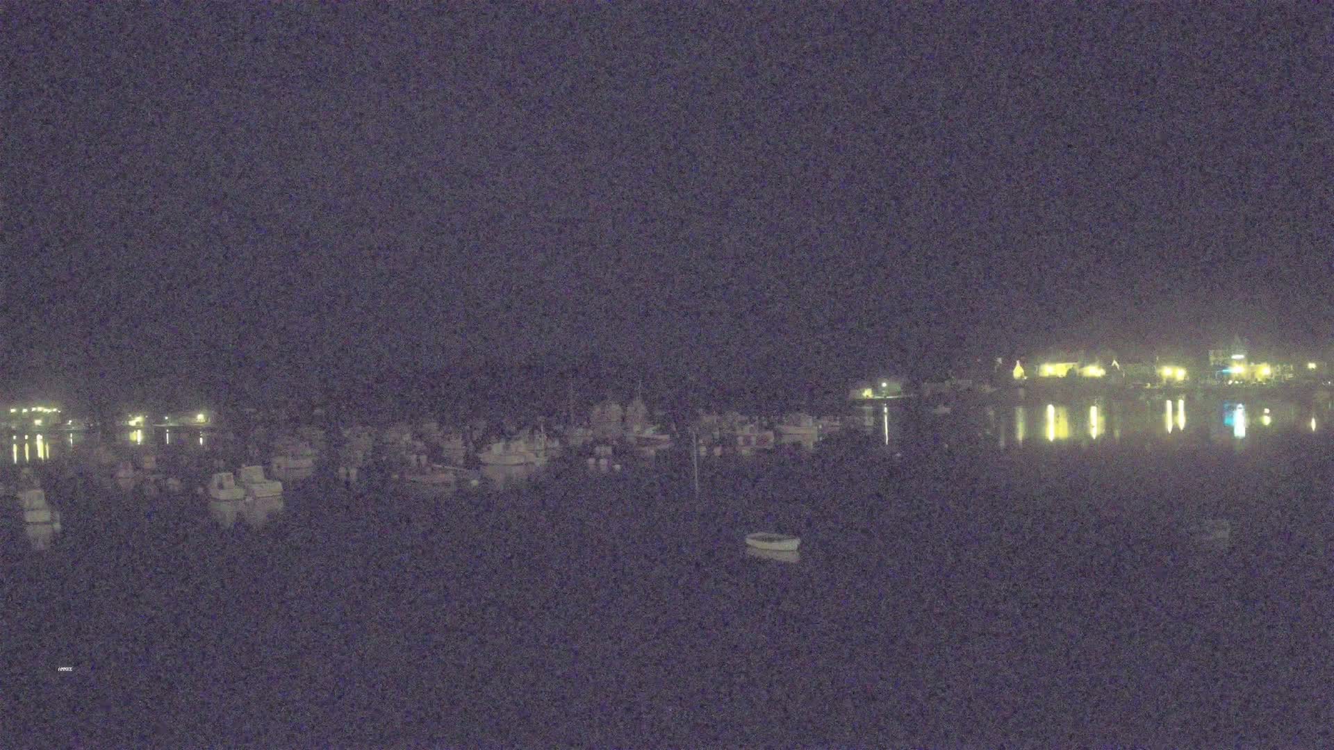 A nighttime view of a calm body of water with numerous boats moored, reflecting the lights from buildings on the opposite shore under a dark sky.