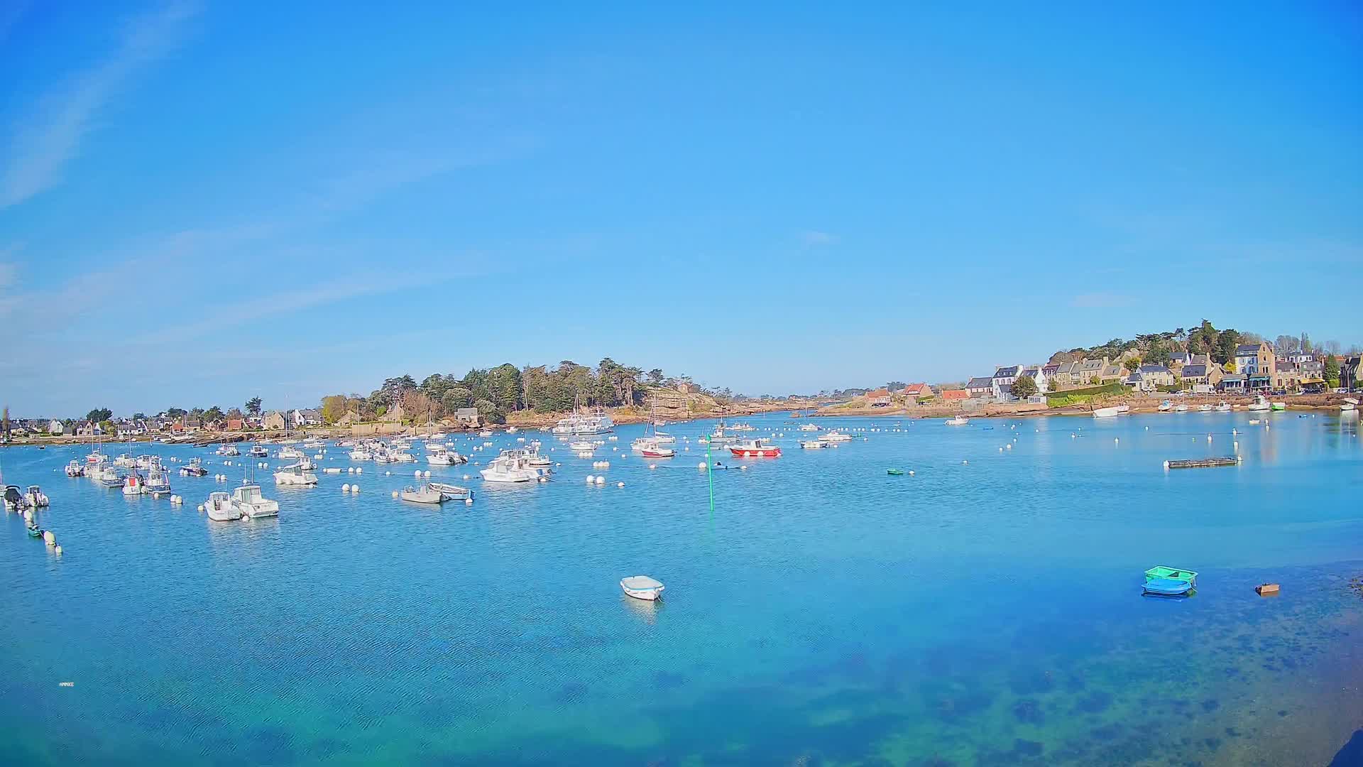 Under a clear blue sky, a calm, turquoise body of water holds numerous small boats near a shoreline village.