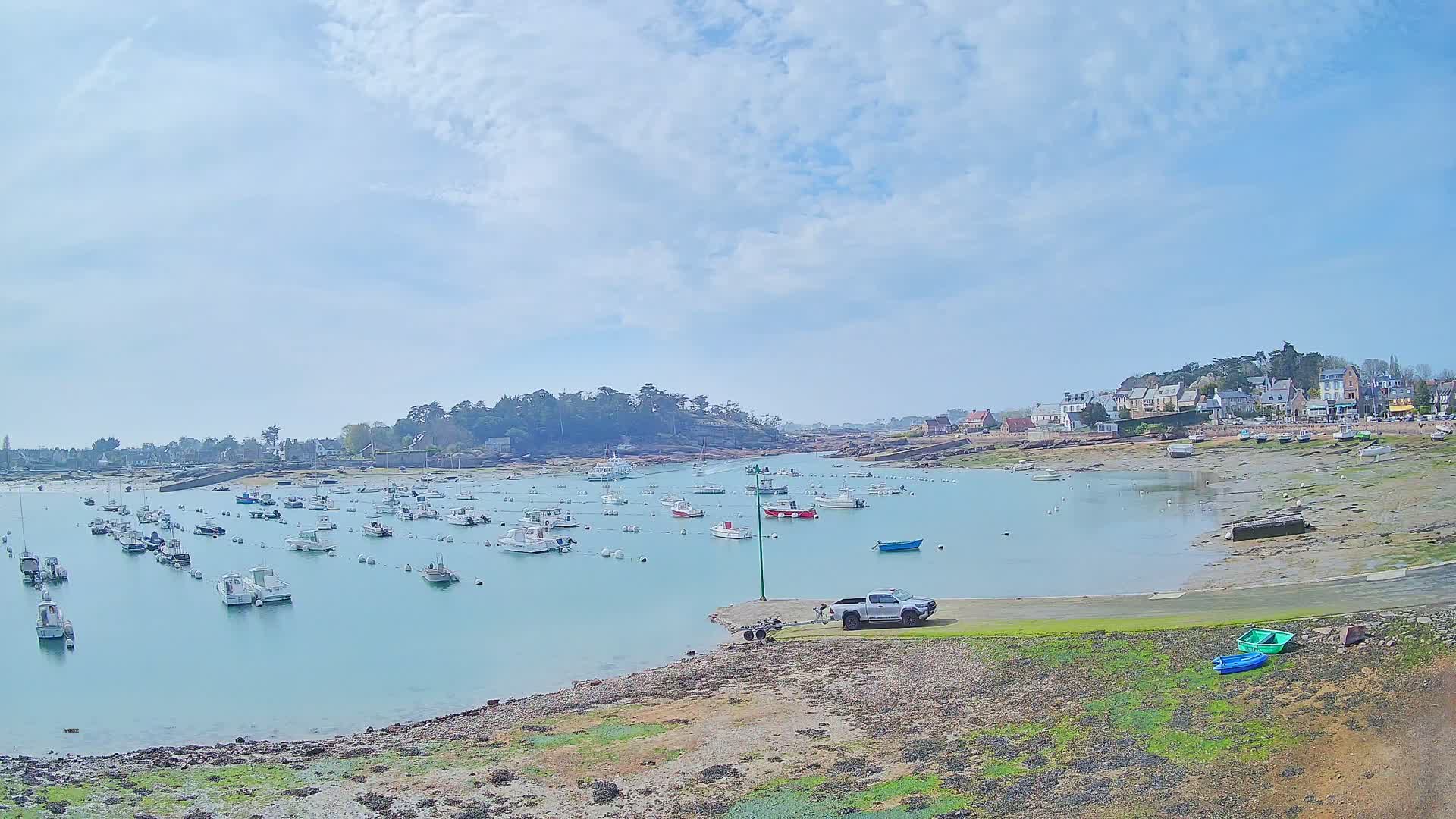 A calm bay with numerous small boats moored, a pickup truck parked near the shore, and a village nestled along the shoreline under a partly cloudy sky.