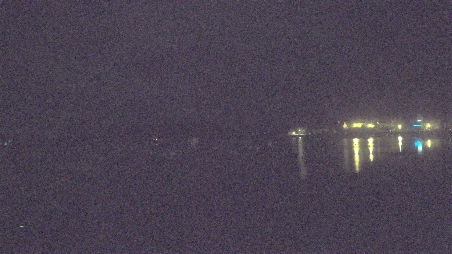 A nighttime view shows a calm body of water reflecting the lights of a distant town under a dark sky.