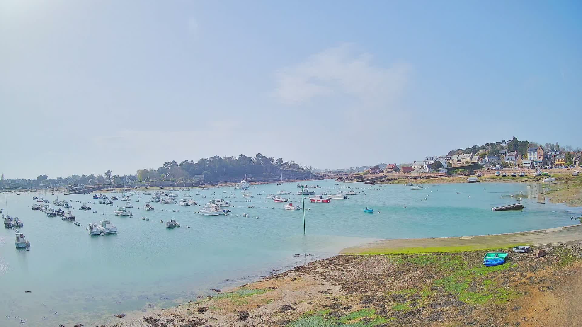 A calm, sunny day shows many small boats moored in a clear, teal harbor next to a small village nestled beside a rocky shoreline.
