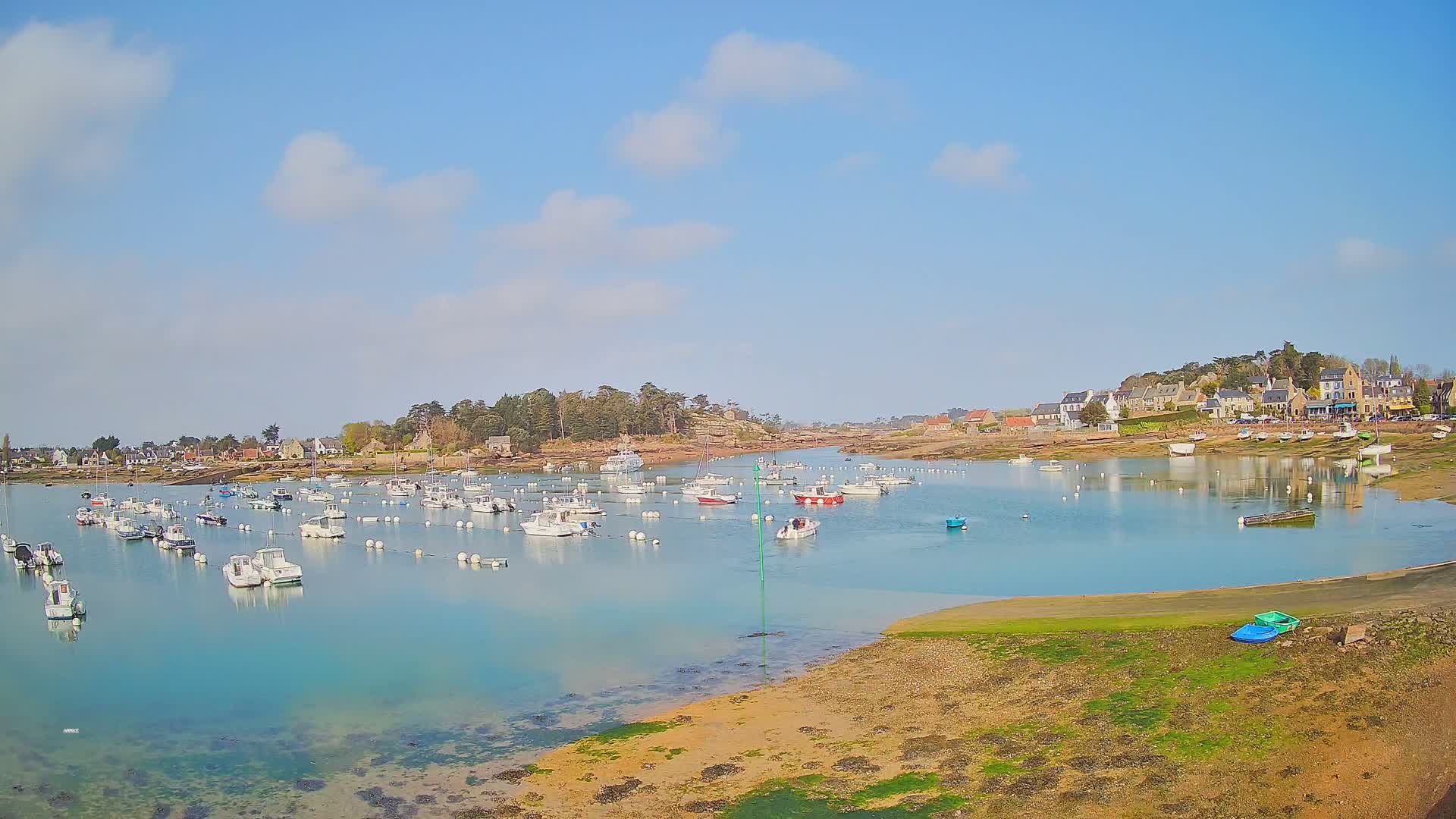 Numerous small boats are moored in a calm, turquoise harbor next to a small village under a mostly sunny sky.