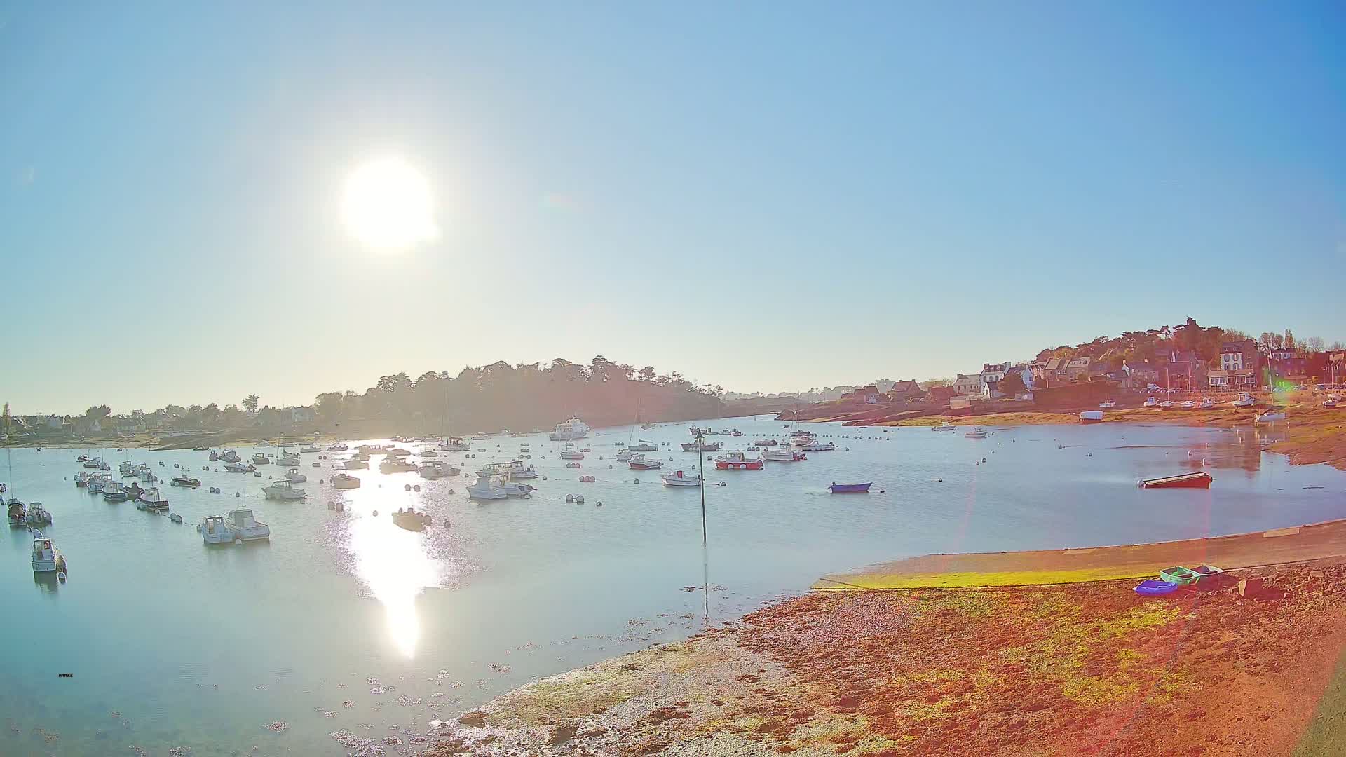 Many boats are moored in a calm bay on a sunny day, with a small village visible on the far shore.