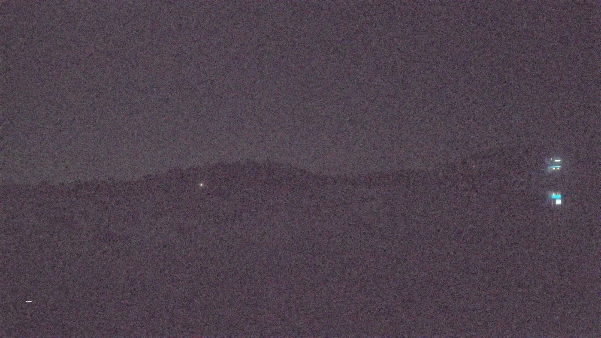 A dark, nighttime landscape shows a distant line of hills under a mostly clear, dark sky, with a few faint lights visible in the distance.