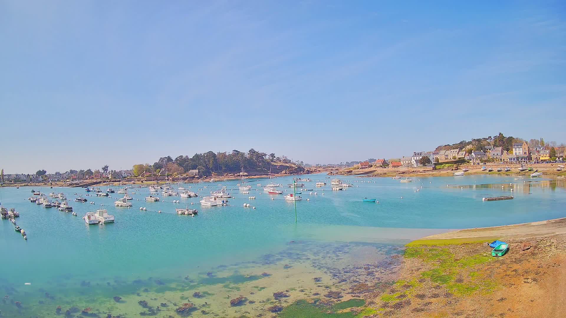 Numerous small boats are moored in a calm, turquoise bay under a clear blue sky, near a shoreline with buildings and trees.