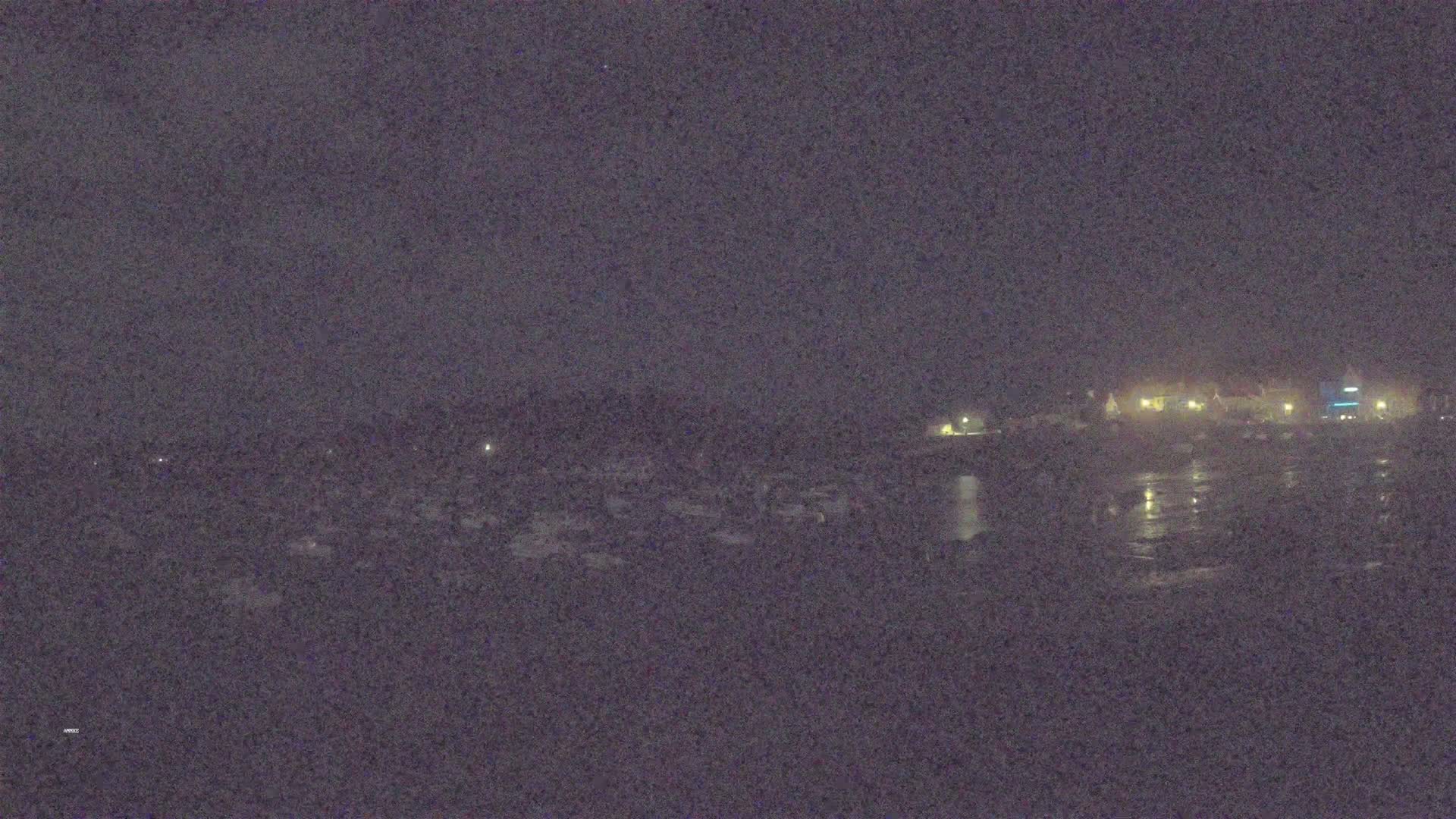 A nighttime view of a dark, possibly wet, expanse of land reflecting faint lights from buildings in the distance under a cloudy sky.