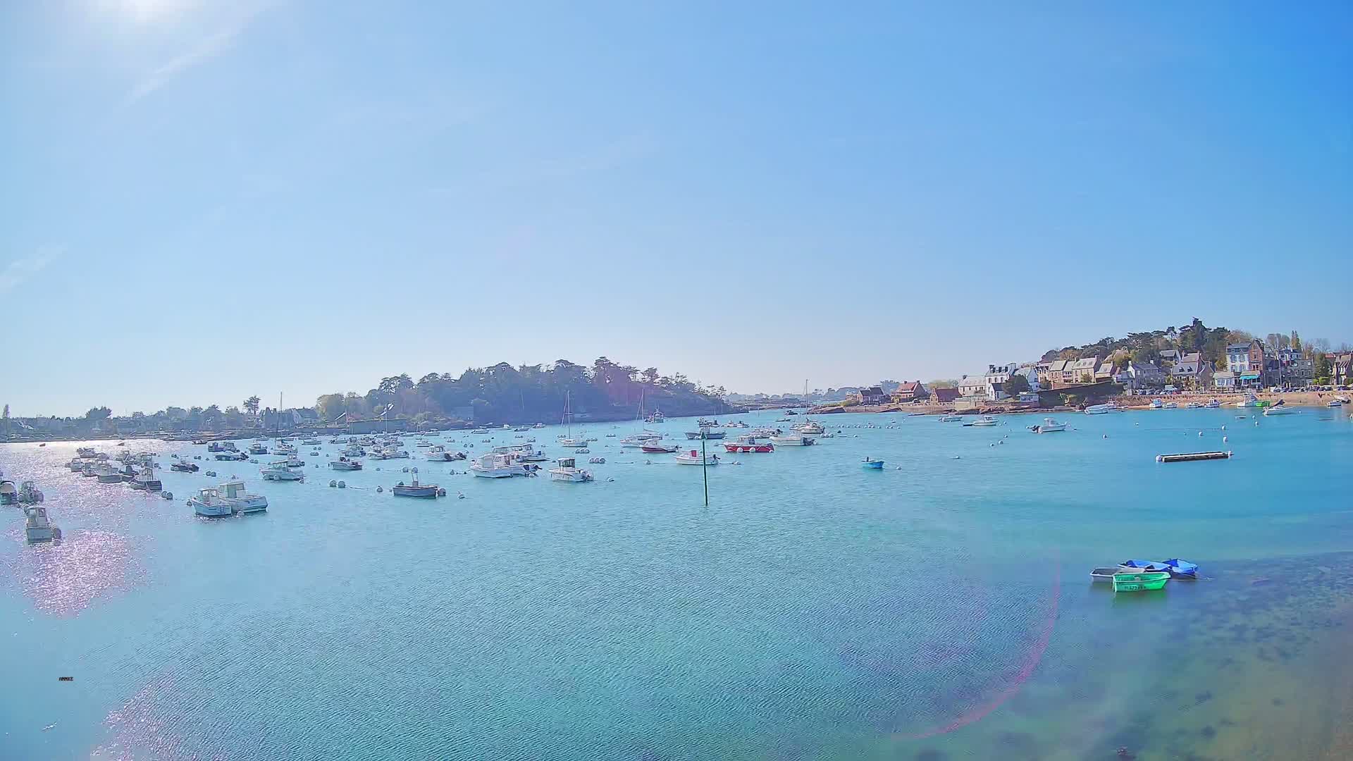 Numerous small boats are moored in a calm, clear aqua-colored bay under a bright, sunny sky, with a small village visible on the far shore.
