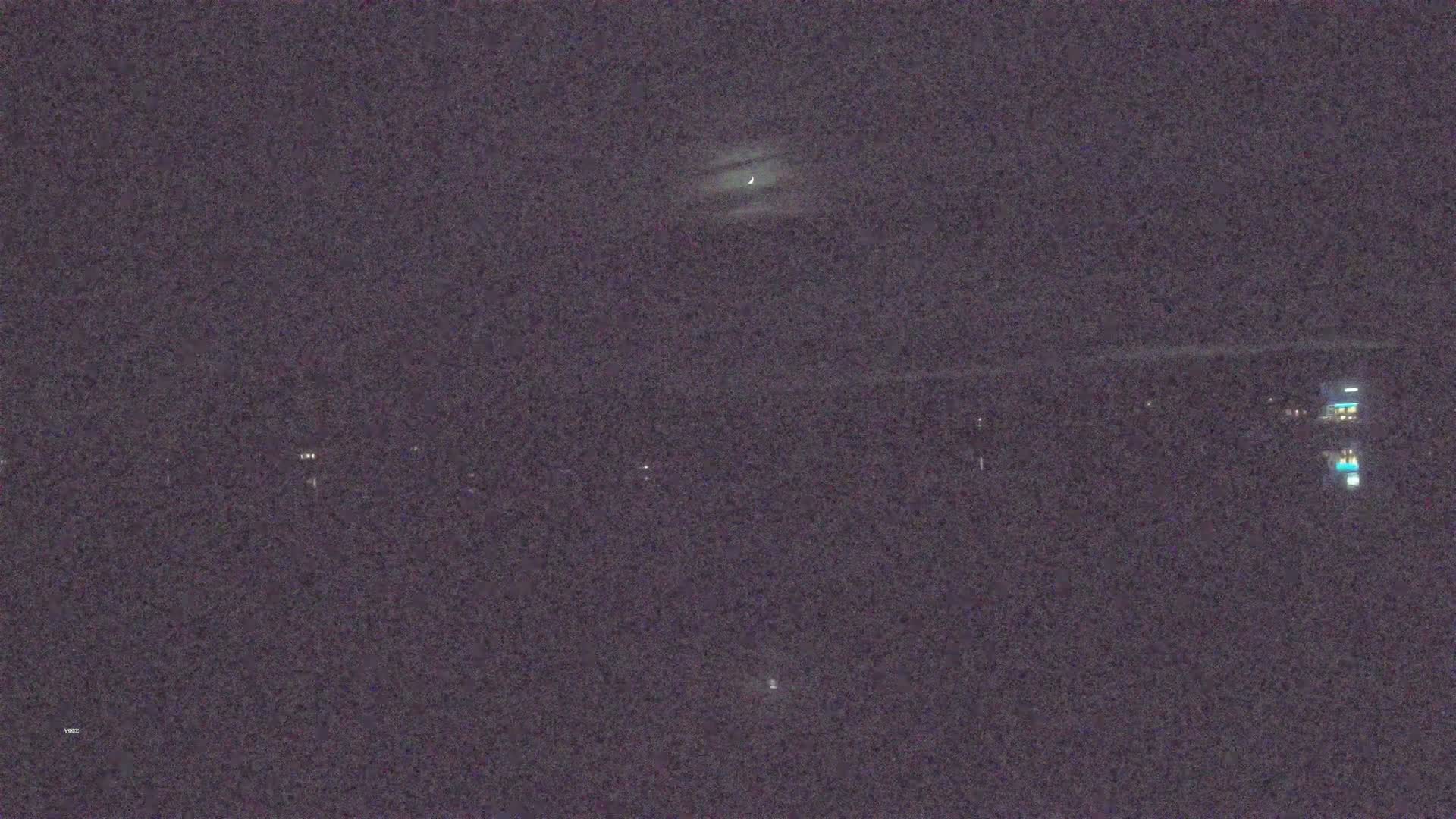A crescent moon is visible in a dark night sky above a distant shoreline with faint lights.