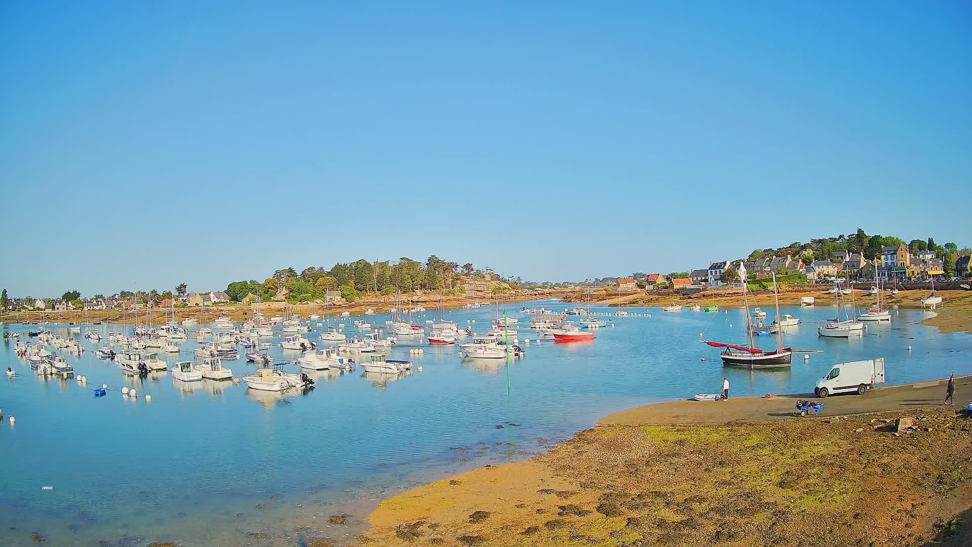 A calm, sunny day reveals a harbor filled with numerous small boats and sailboats, nestled beside a shoreline village and a rocky outcrop.