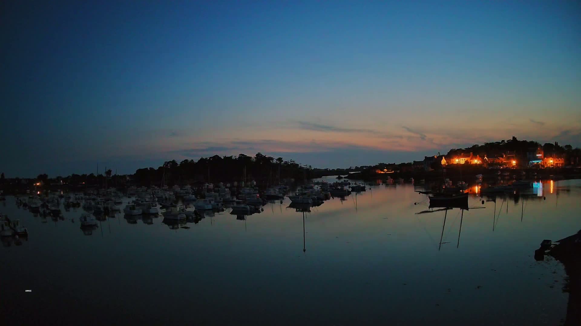 Many boats are moored in a calm harbor at twilight, with a small town's lights visible on the far shore.