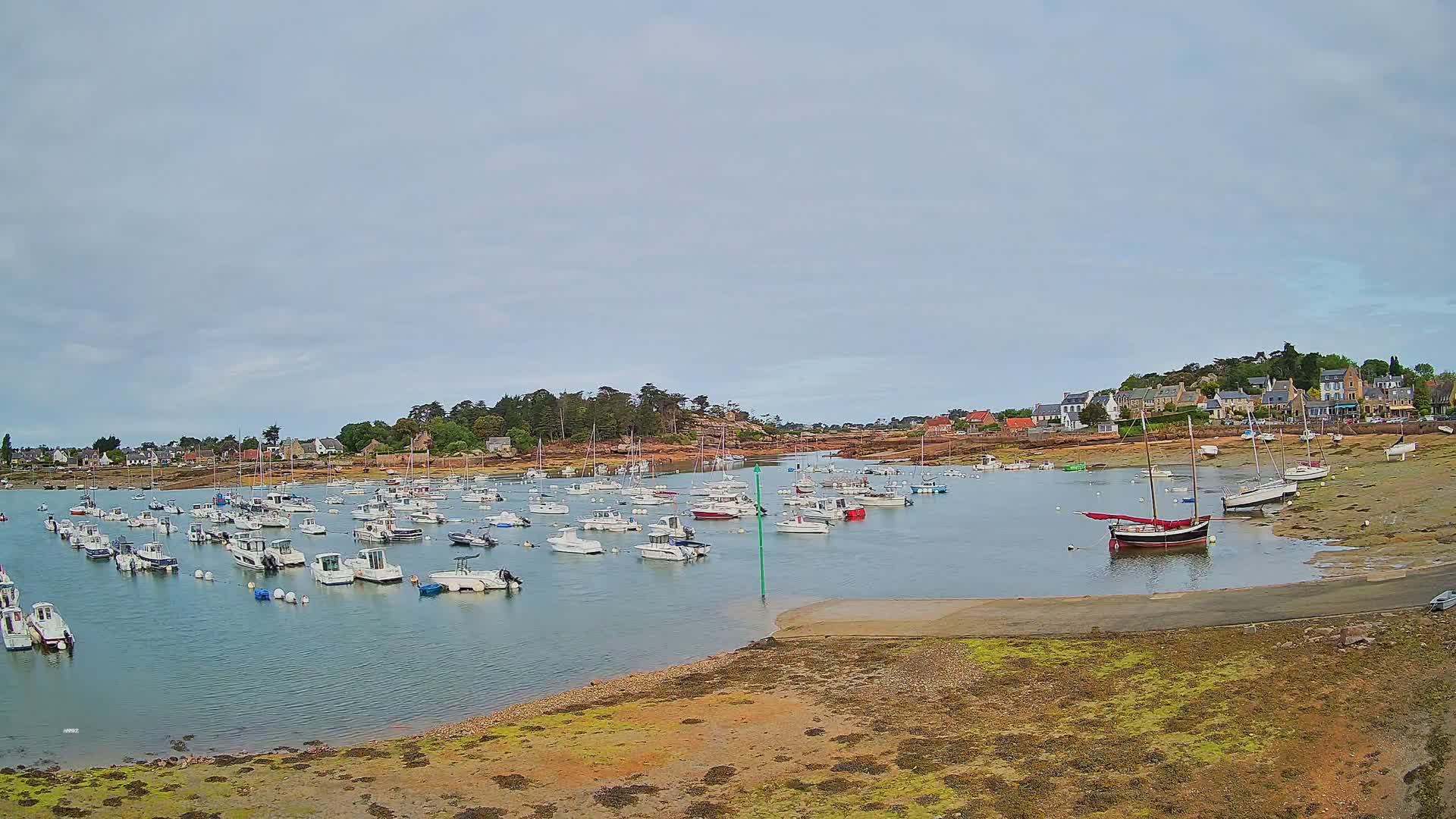 Many small boats are moored in a calm bay near a small village under an overcast sky.