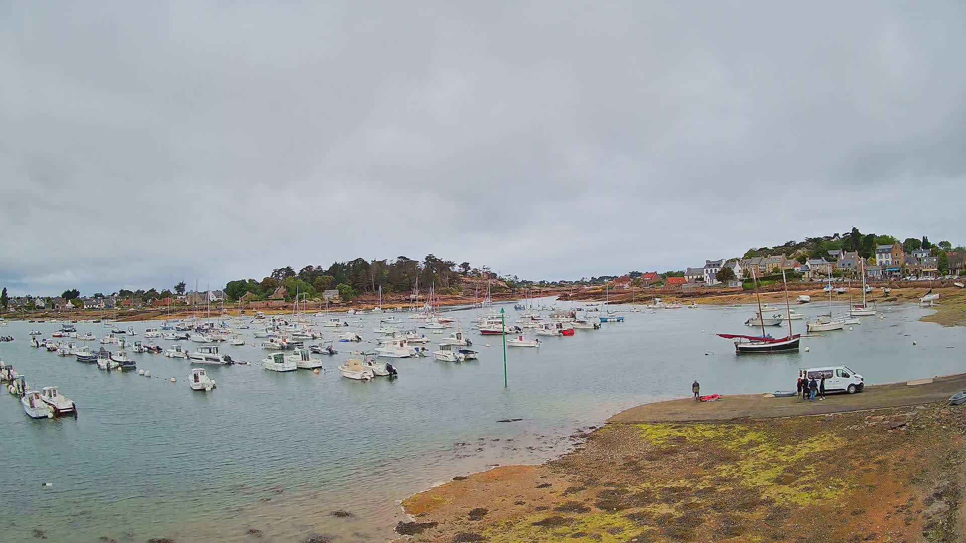 Many small boats are moored in a calm harbor under an overcast sky, with a small village visible on the shore.