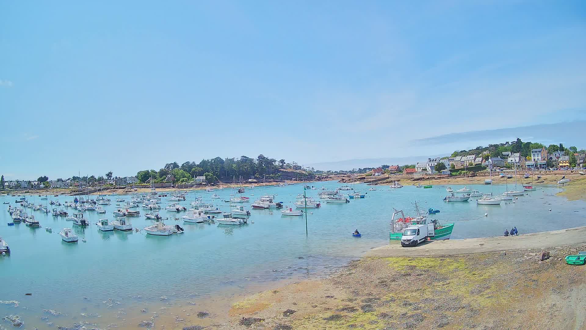 A calm, sunny day reveals a harbor filled with numerous small boats, nestled beside a small village situated on a rocky coast.