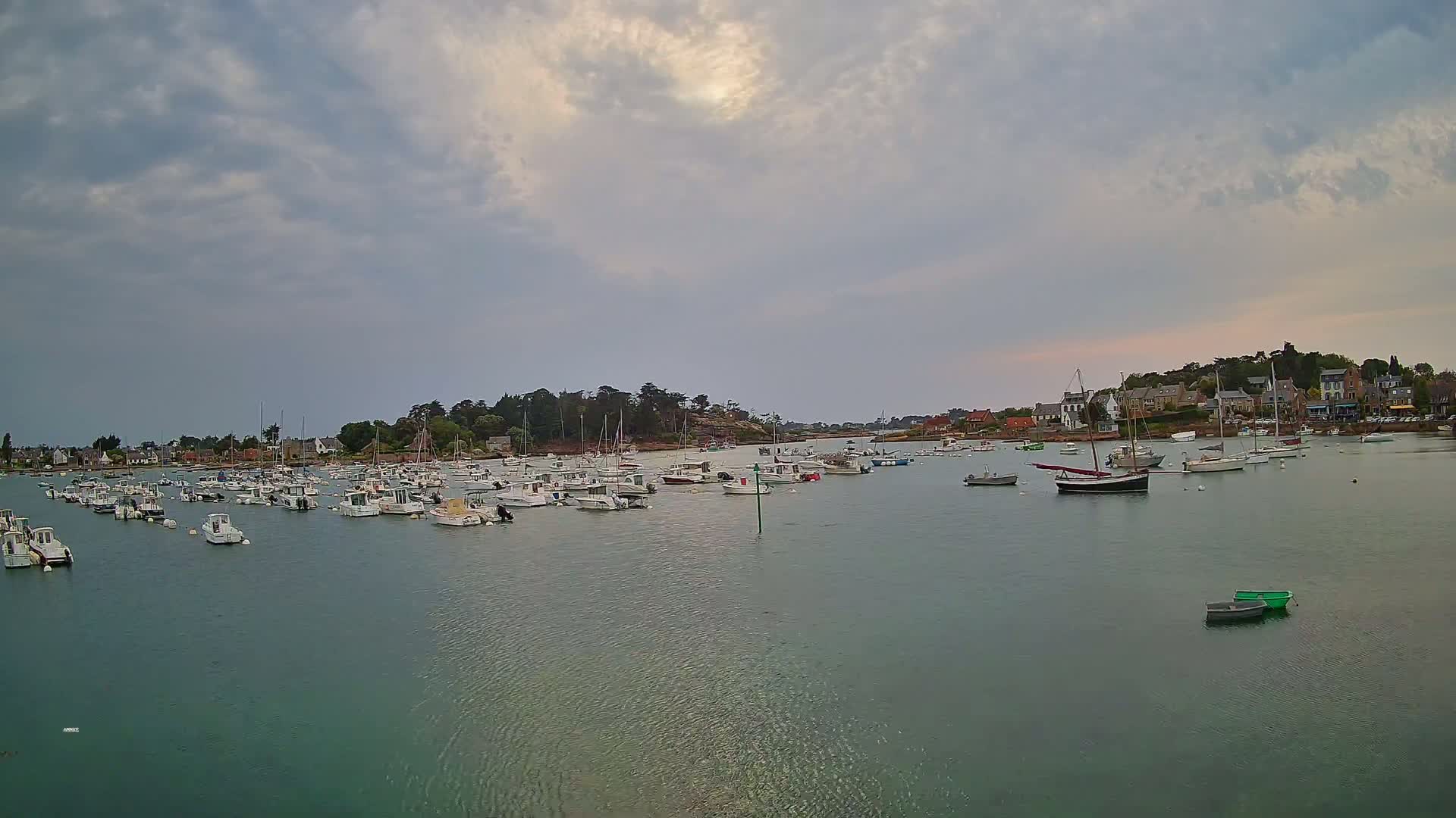 Numerous boats are moored in a calm harbor under a cloudy sky, with a small village visible on the far shore.