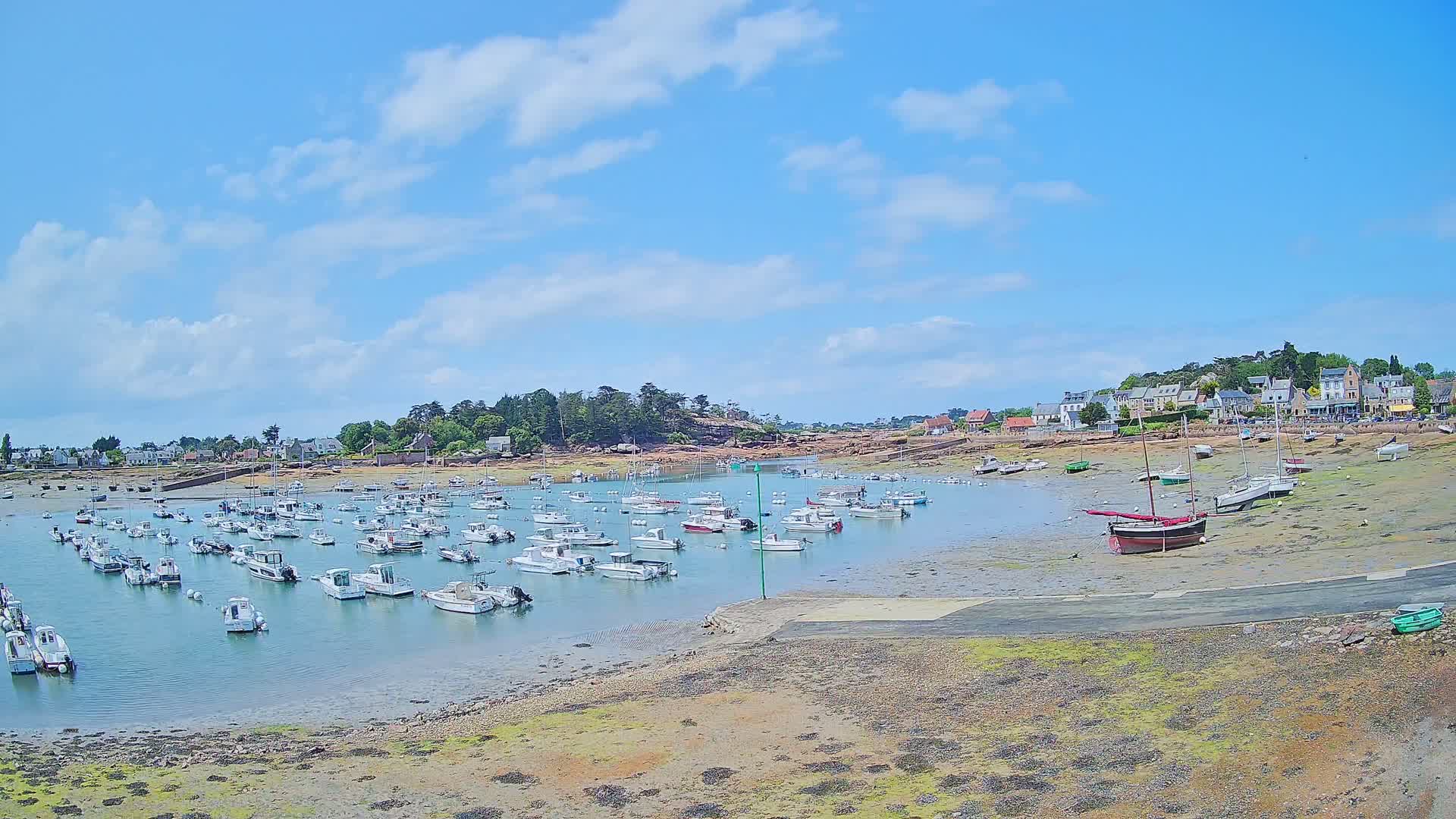 Numerous small boats are moored in a calm harbor on a sunny day with partly cloudy skies, near a small village nestled amongst greenery.
