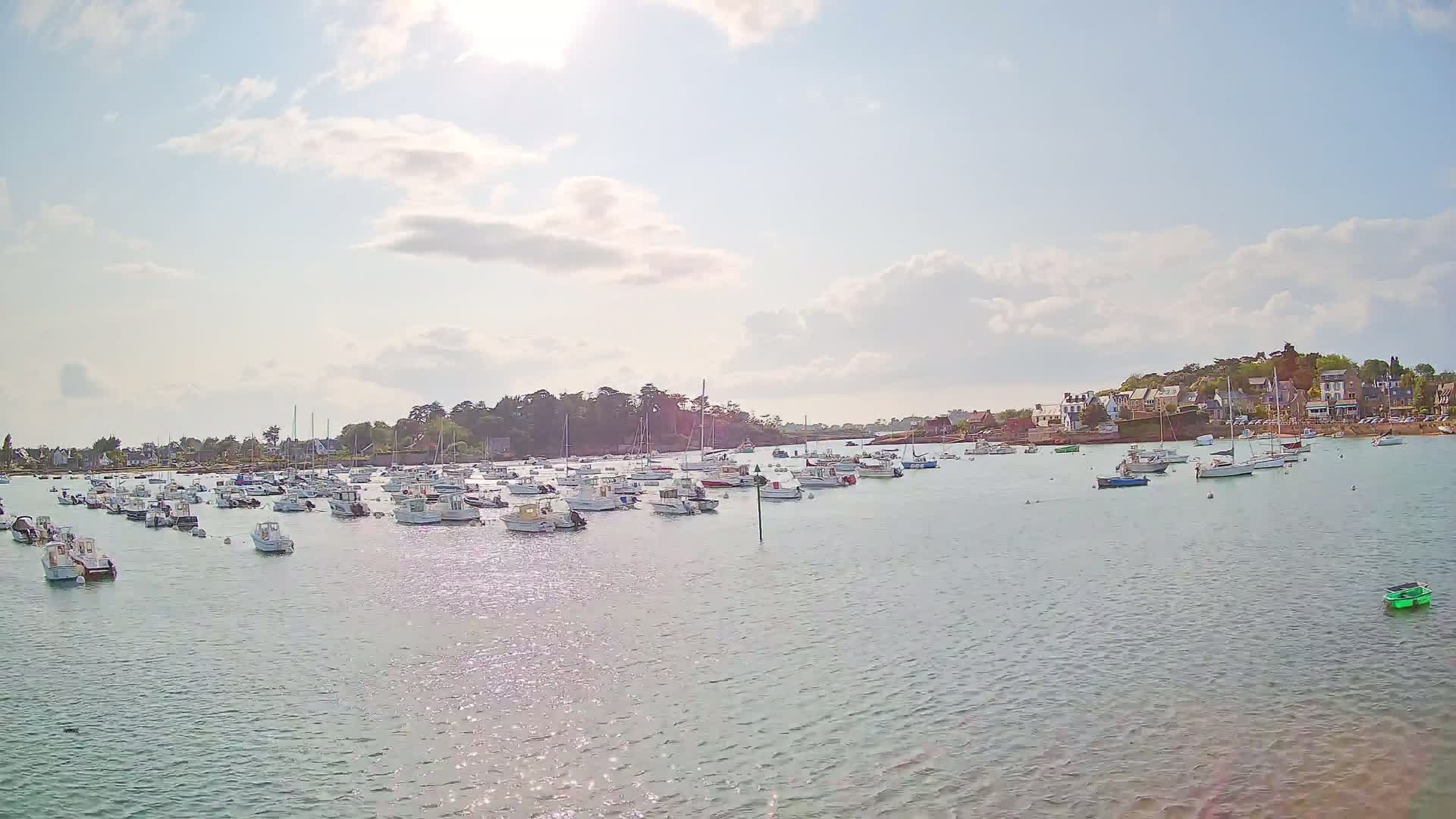 Many small boats are moored in a calm harbor under a partly sunny sky, with a village visible in the background.