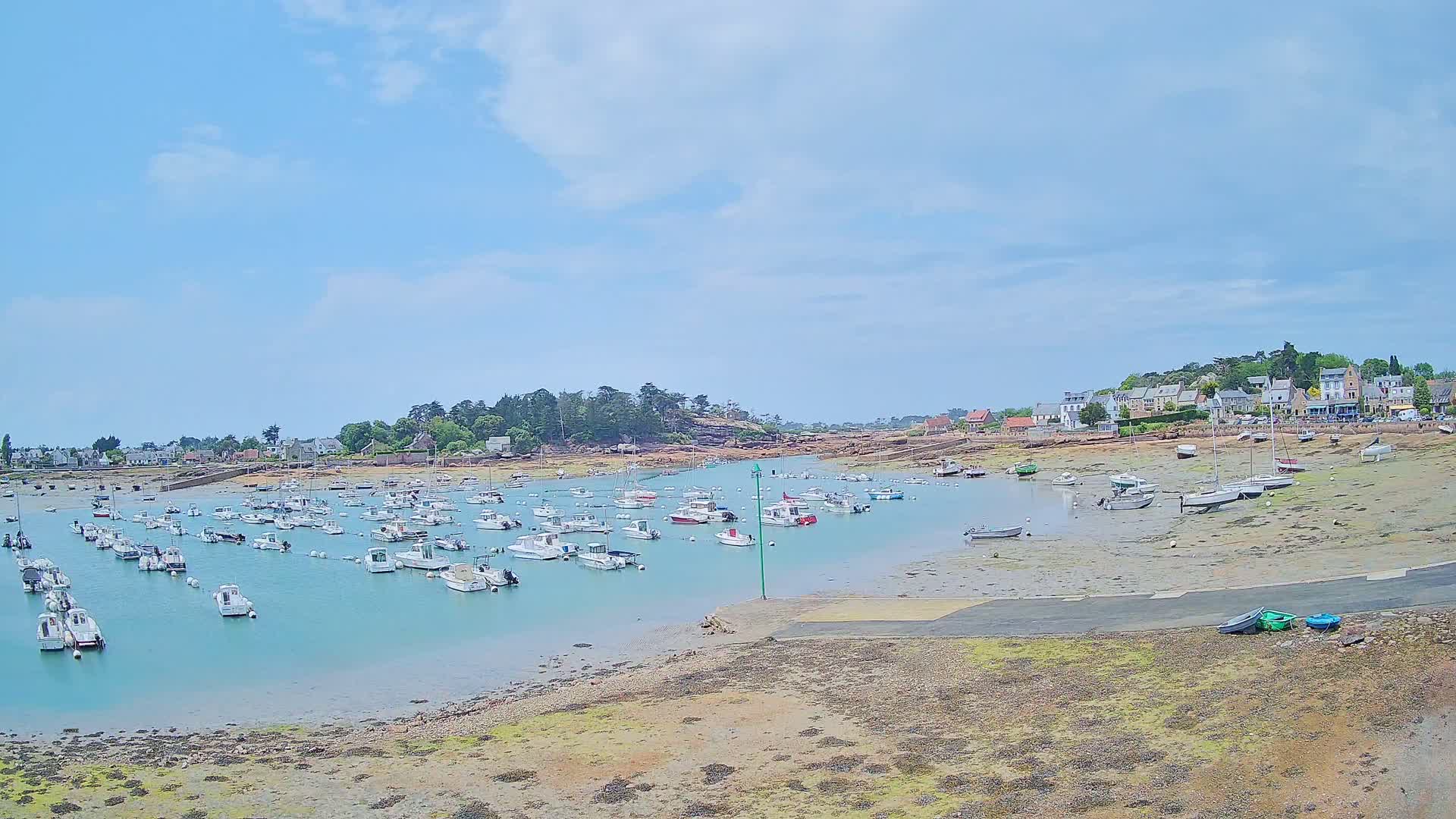 A calm, sunny day reveals a harbor filled with numerous small boats, some in the water and others resting on exposed mudflats, with a small village nestled on the shore behind.