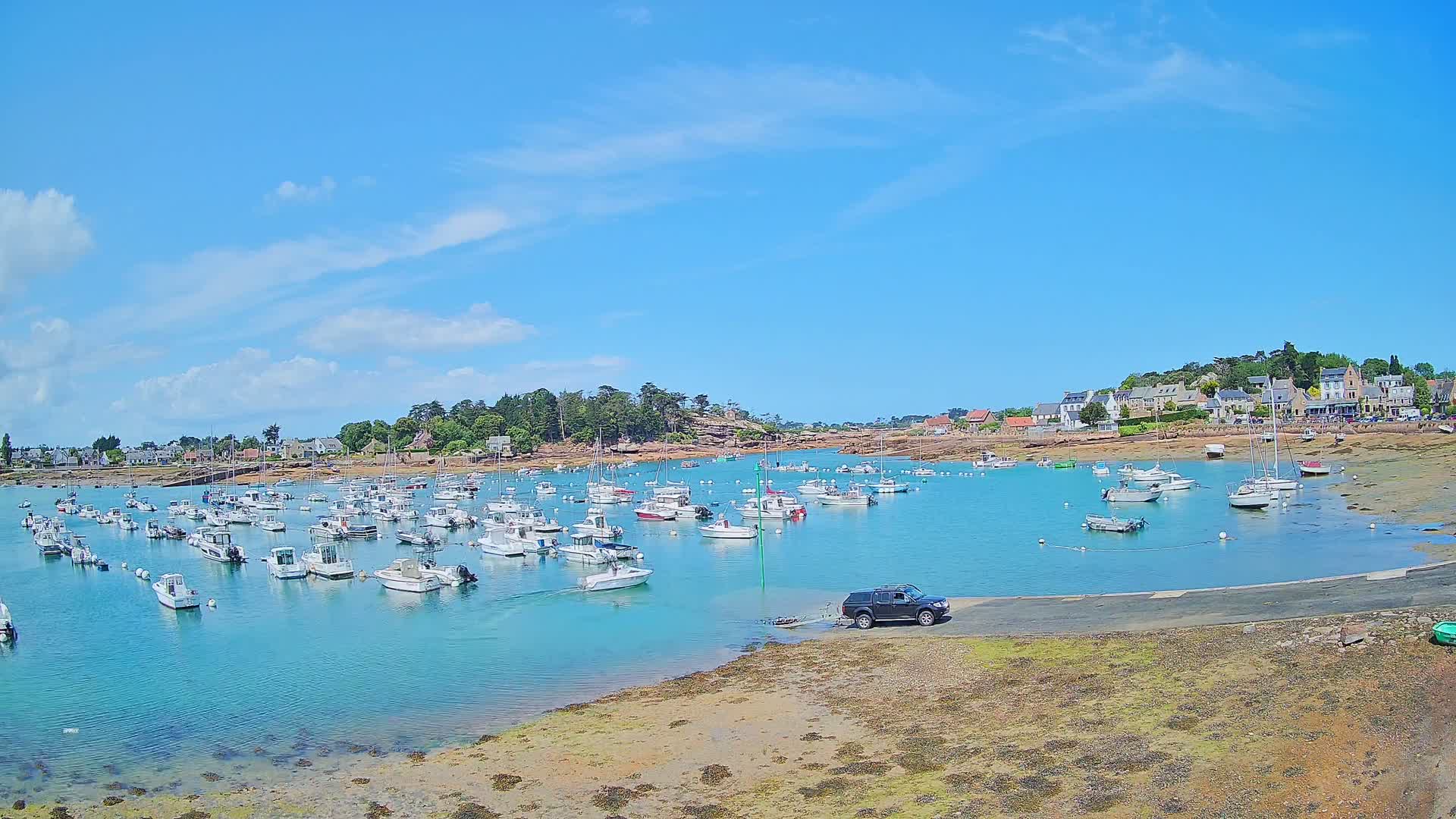 A calm, sunny day reveals a harbor filled with numerous boats, a small town nestled on a rocky shoreline, and a dark-colored SUV parked near the water's edge.