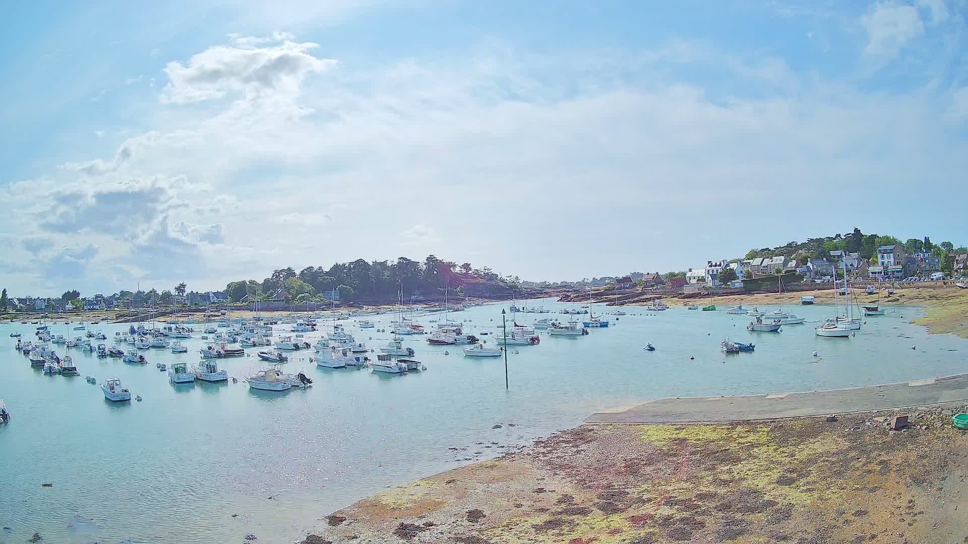 Under a partly cloudy sky, numerous small white boats are moored in a calm bay next to a small village.