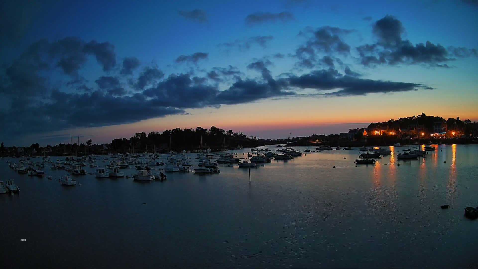 Under a partly cloudy sky, numerous small white boats are moored in a calm bay next to a small village.