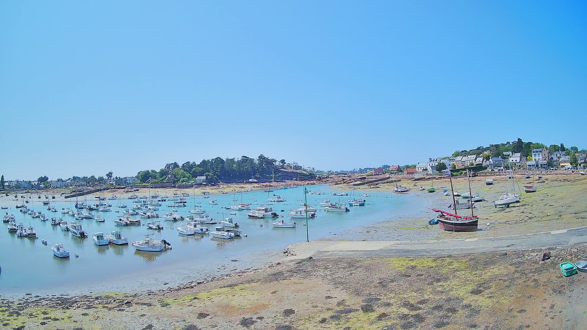 A calm, sunny day reveals a harbor filled with numerous small boats, some grounded on exposed mudflats, near a small village.