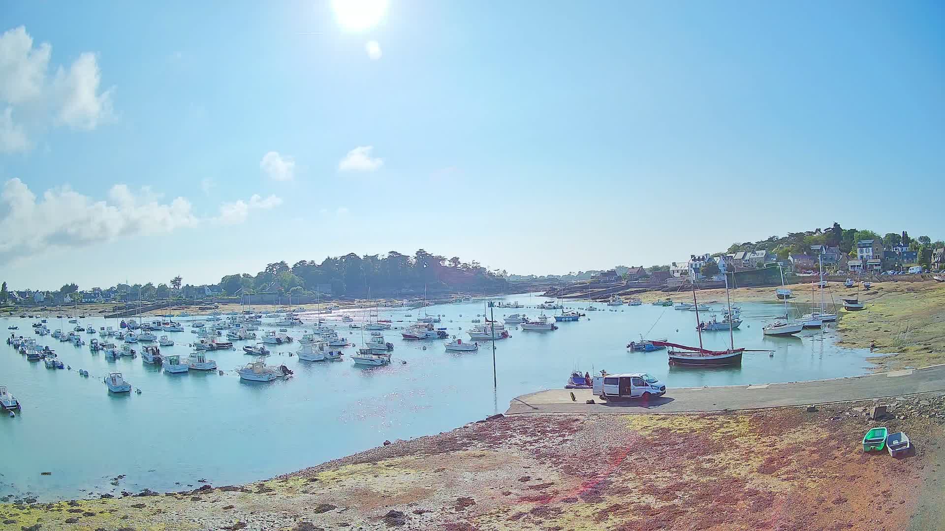 A sunny day reveals a calm harbor filled with numerous small boats, a van parked nearby, and a small village nestled on the shore.
