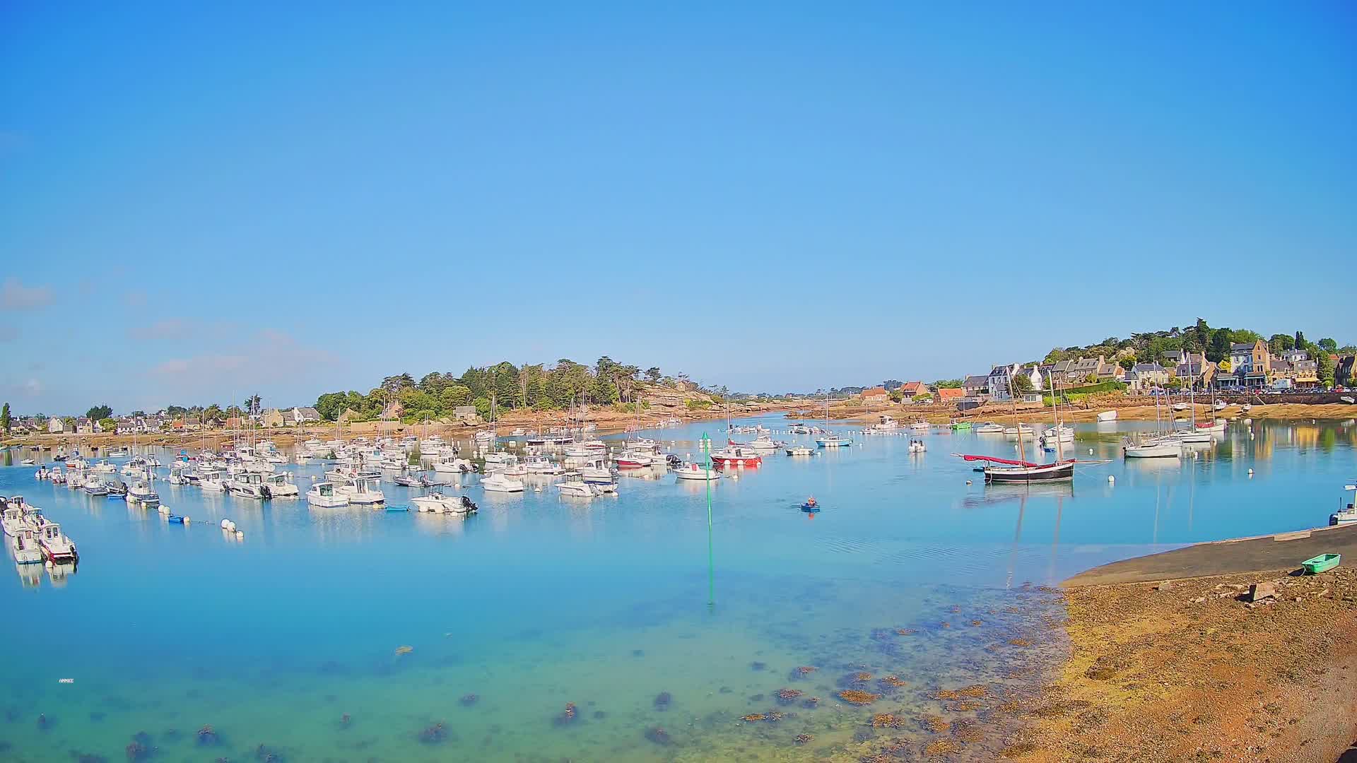 A calm, sunny day reveals a harbor filled with numerous small boats, nestled alongside a shoreline with low-lying buildings and green vegetation.