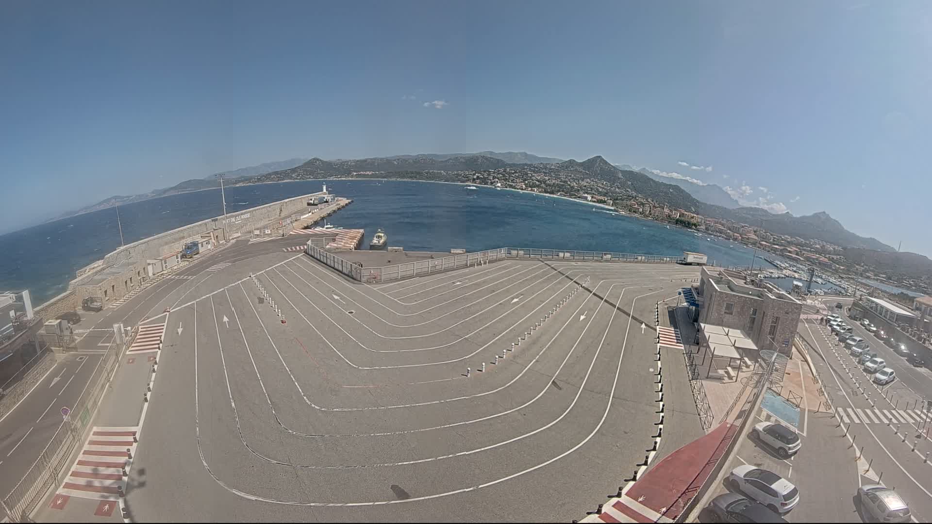 A large, empty ferry terminal with marked lanes curves down to a harbor, under a bright, sunny sky overlooking a calm sea and distant mountains.