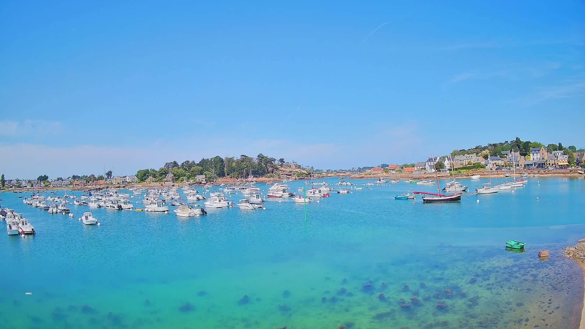 A calm, clear blue body of water filled with numerous small boats is nestled in a coastal town under a bright, sunny sky.