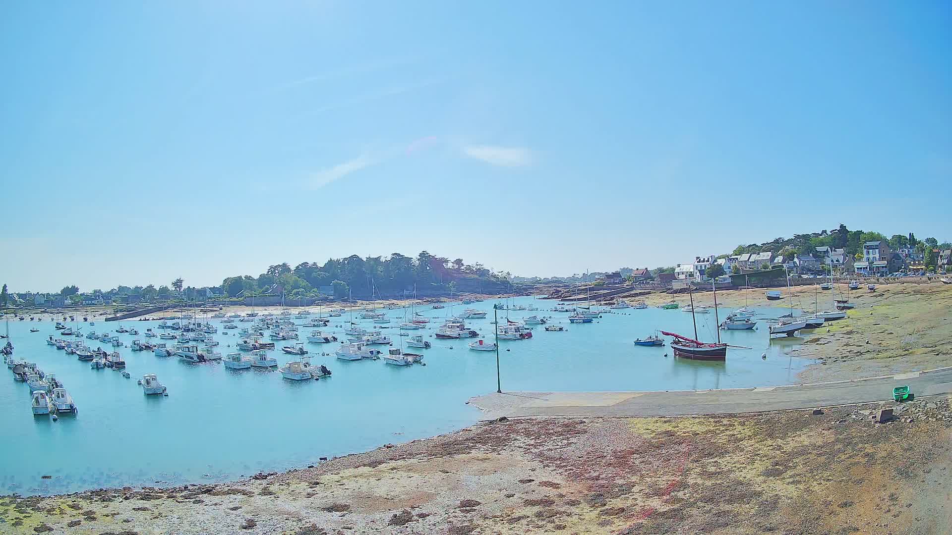 Many small white boats are moored in a calm, turquoise harbor on a sunny day, with a small village and low-lying land visible in the background.