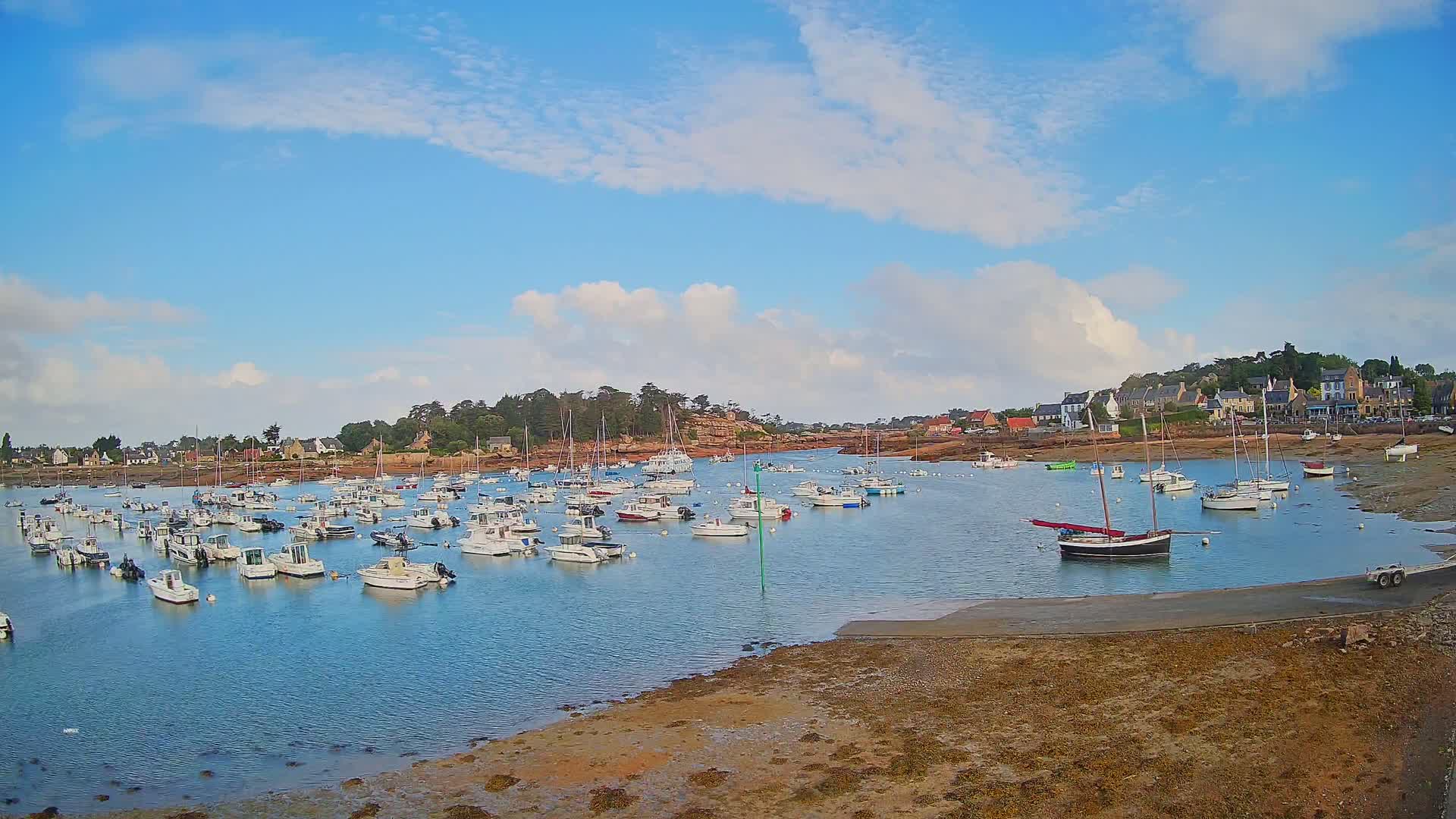 A calm harbor filled with numerous boats is nestled beside a small town under a mostly sunny sky with scattered clouds.