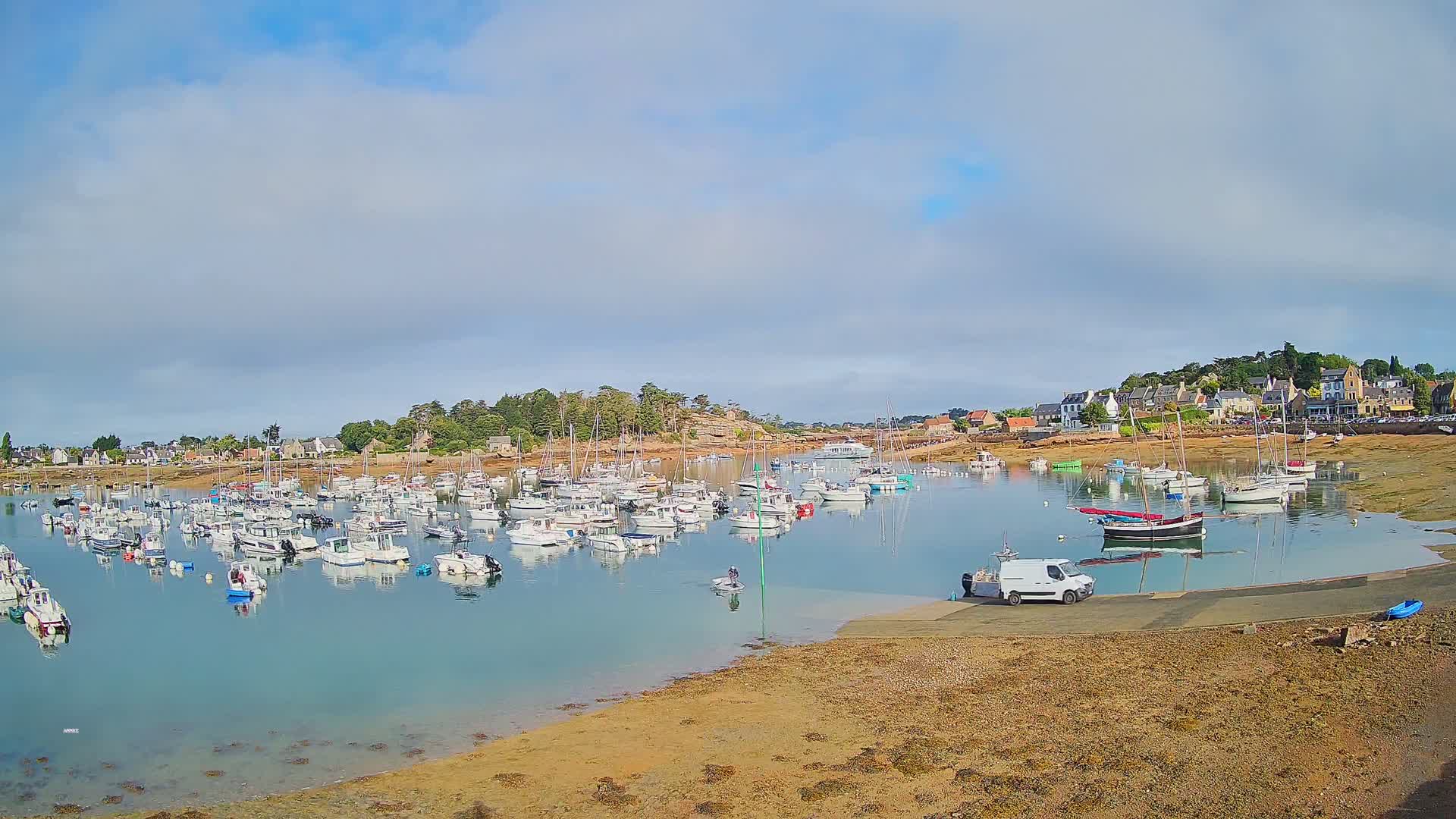 A calm harbor filled with numerous sailboats and motorboats under a partly cloudy sky, with a small town and a sandy shore visible in the background.