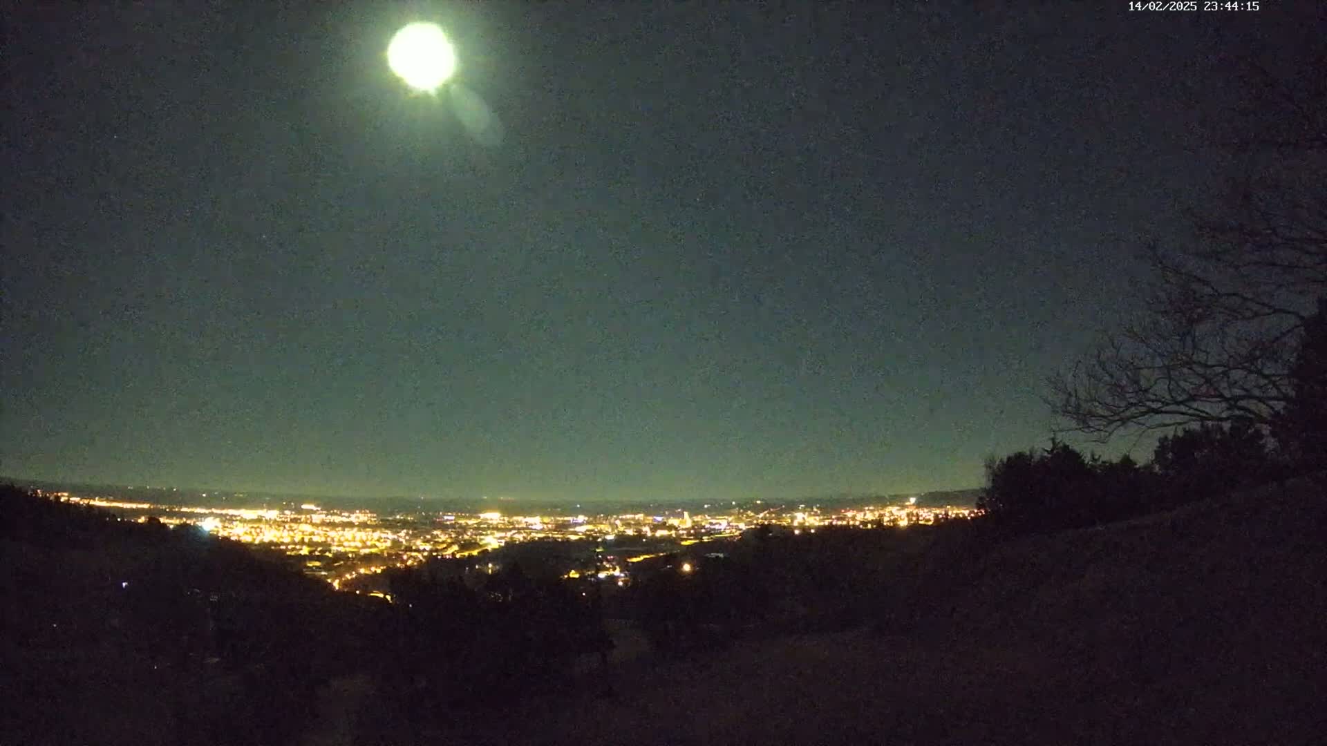 A brightly lit city is visible in the distance under a mostly clear night sky with a very bright moon.