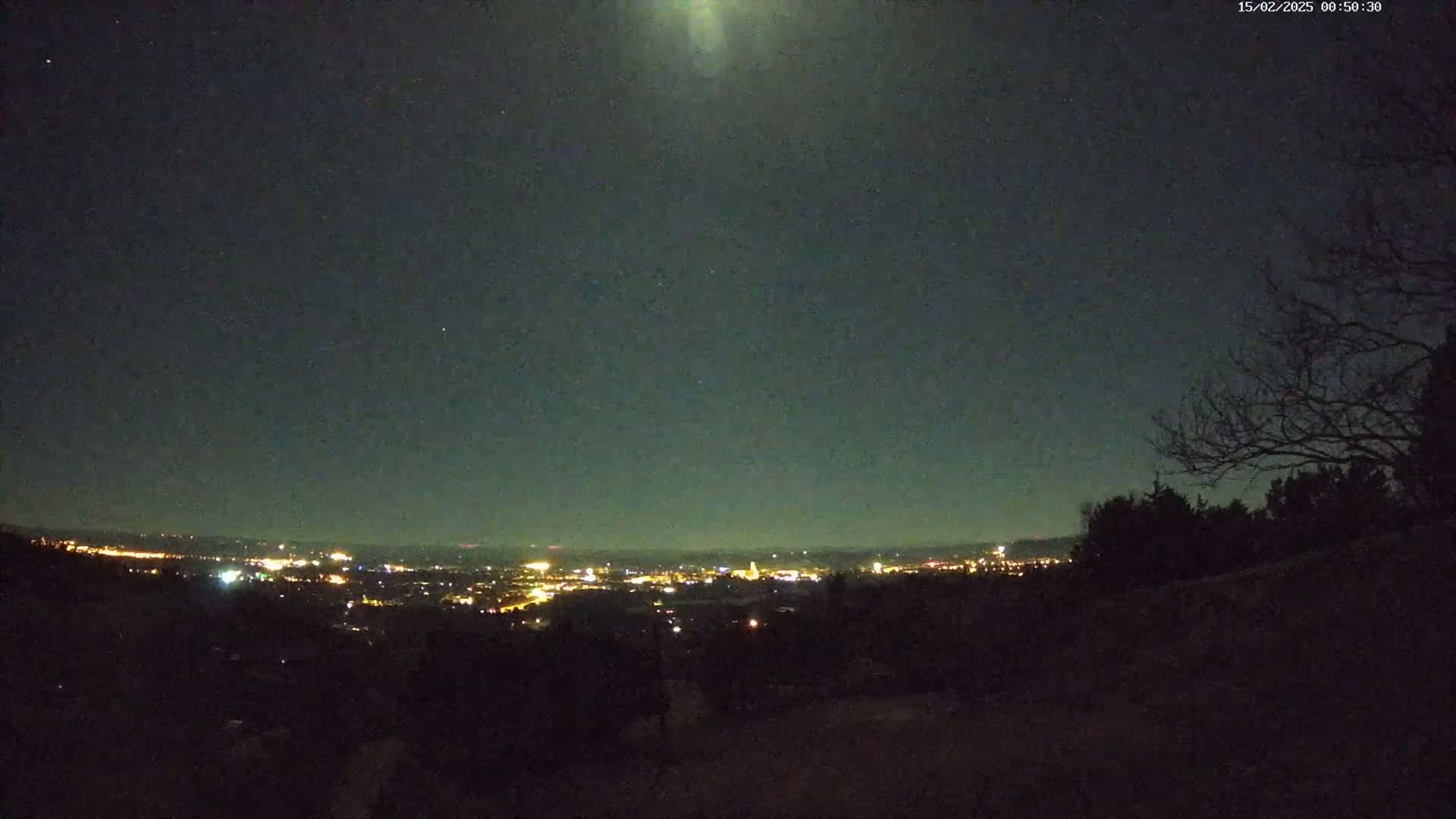 A nighttime view of a city's lights on the horizon from a hilltop, under a mostly clear, dark sky with a bright moon or light source visible.