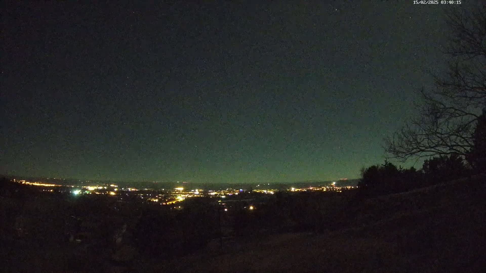 A nighttime view from a hilltop shows a distant city illuminated with lights under a clear, dark sky dotted with stars.